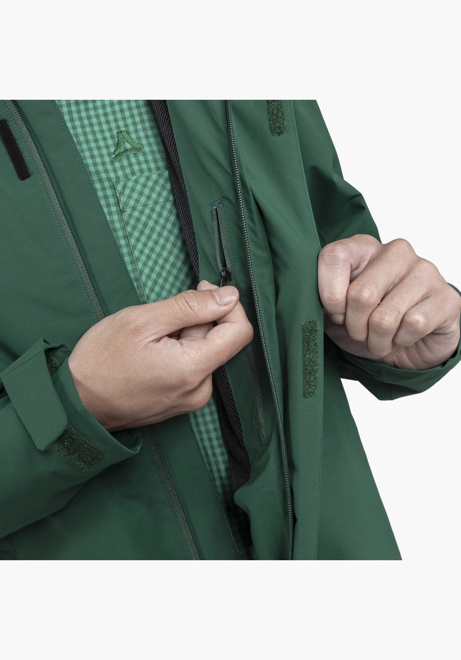Jacket Gmund M, detail view of the green fabric and the inner zip pocket being opened by a hand, alongside hook-and-loop fasteners on the flap and the visible Schöffel-Logo on the shirt underneath, close-up shot against a white/neutral background.