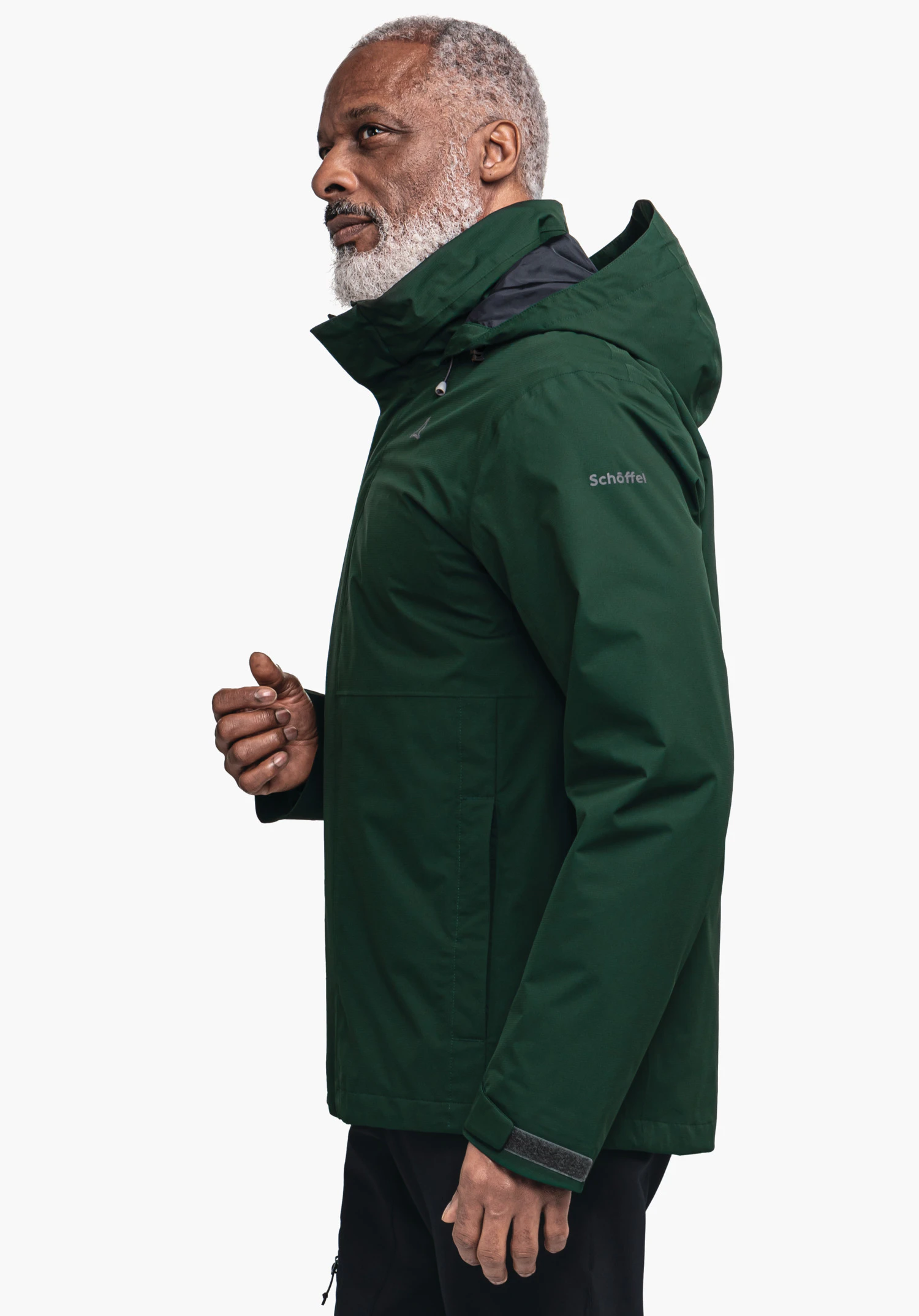 Jacket Gmund M veste vert foncé portée par un homme à barbe grise, debout de profil, capuche relevée, devant un fond blanc neutre. Sur la manche: "Schöffel".