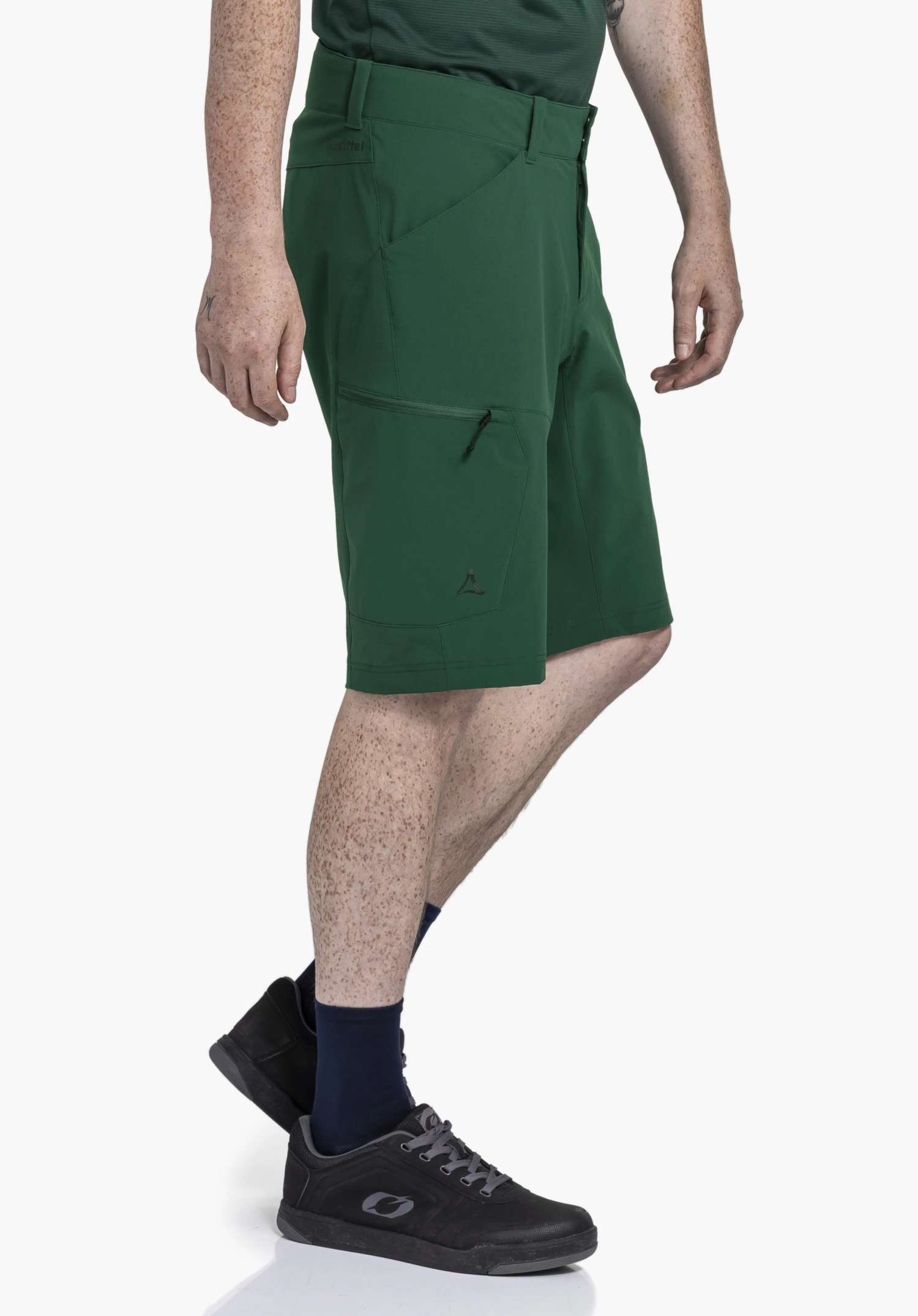 Shorts Danube M in Grün: Das Modell steht seitlich vor einem neutralen Hintergrund und präsentiert die Passform der Hose. Das dezente Schöffel-Logo ist am unteren Beinabschluss sowie dezent am Bund sichtbar.