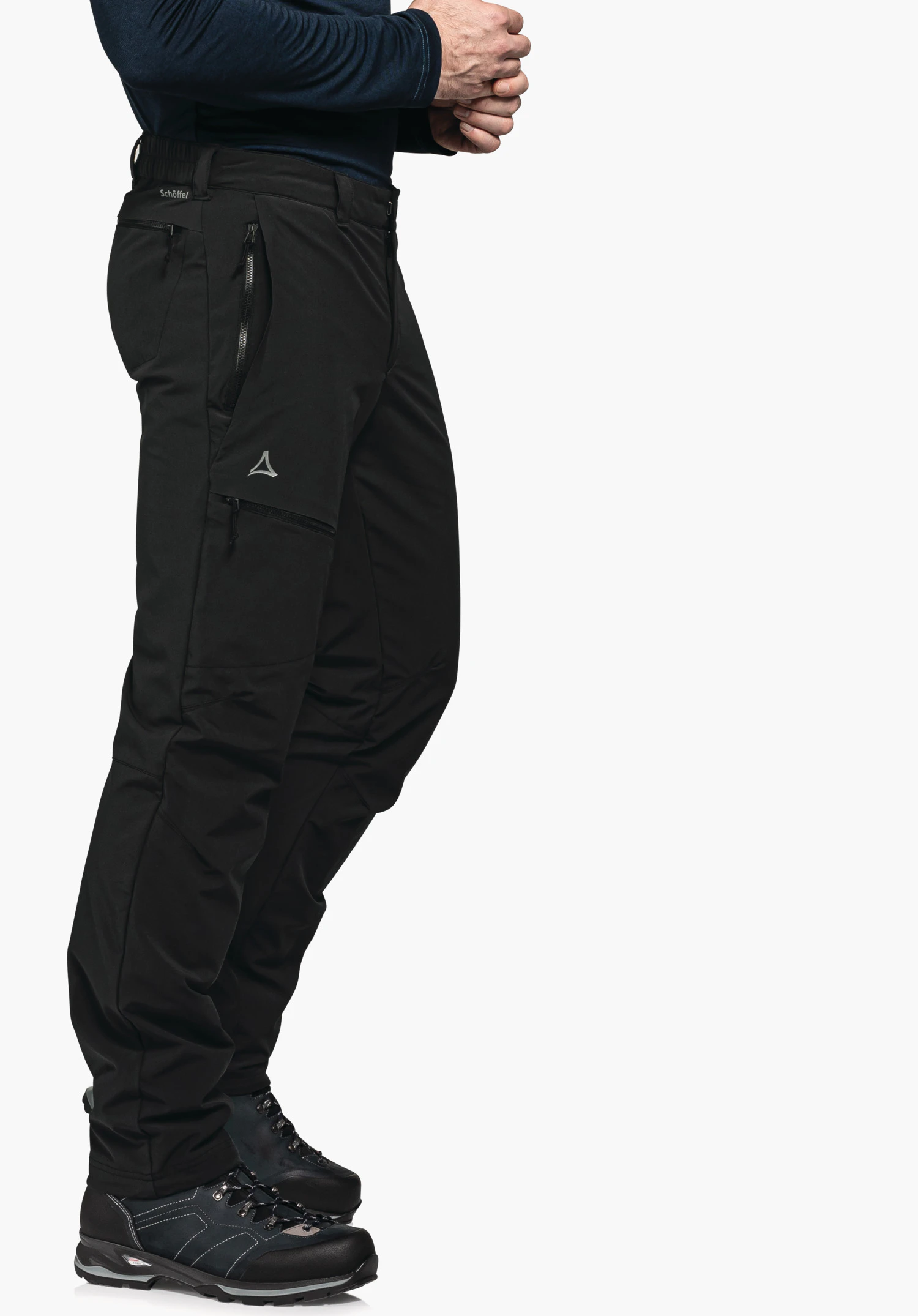 Pants Koper1 Warm M black outdoor trousers, side view with slightly bent legs; person with folded hands wears a dark blue top; white studio background. Schöffel logo visible; text: "Schöffel".