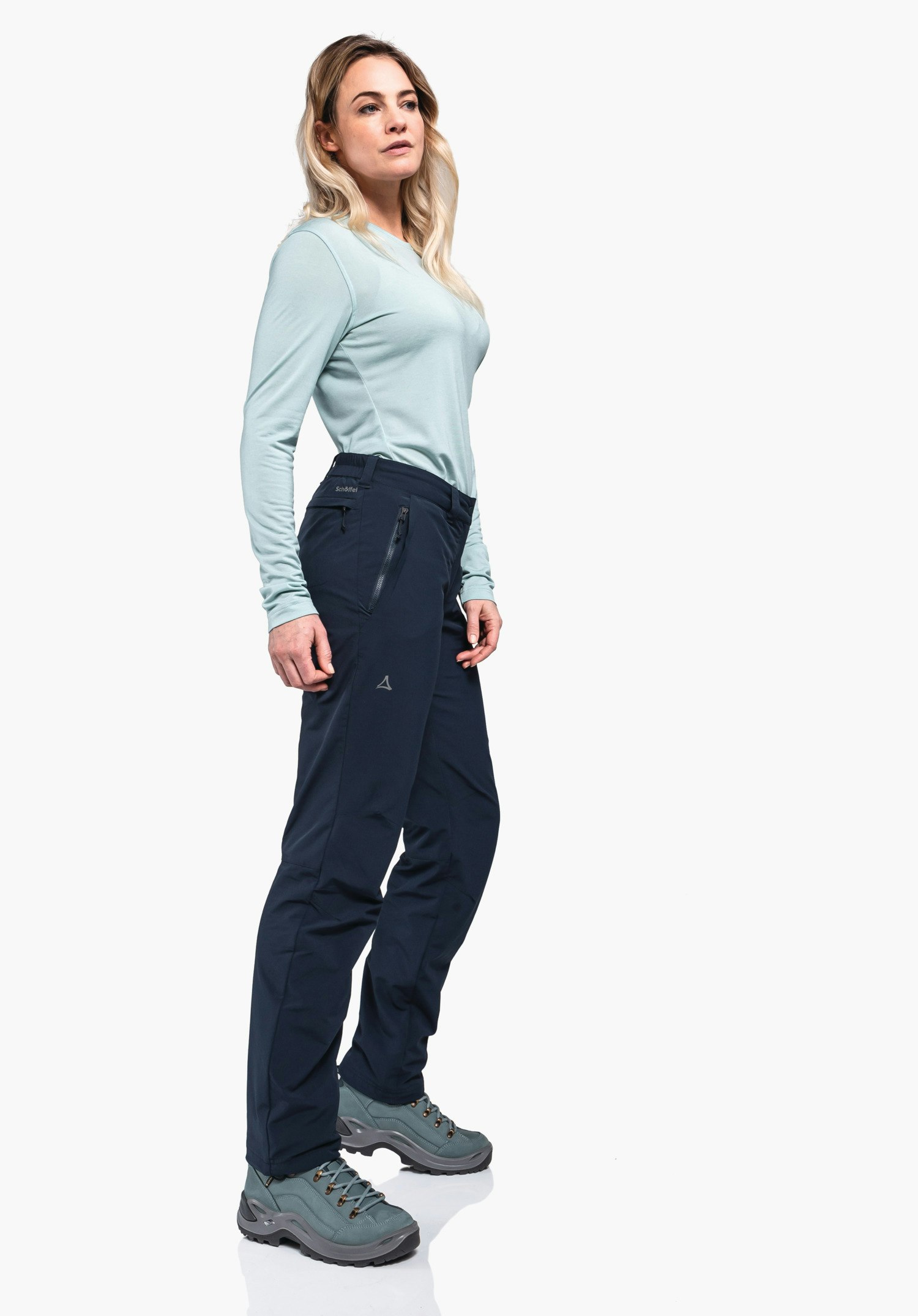 Pants Engadin1 Warm L dark blue hiking trousers with Schöffel logo; woman stands side-on with relaxed hands, wearing a light blue shirt and light green hiking shoes in front of a neutral studio background. Text: "Schöffel".
