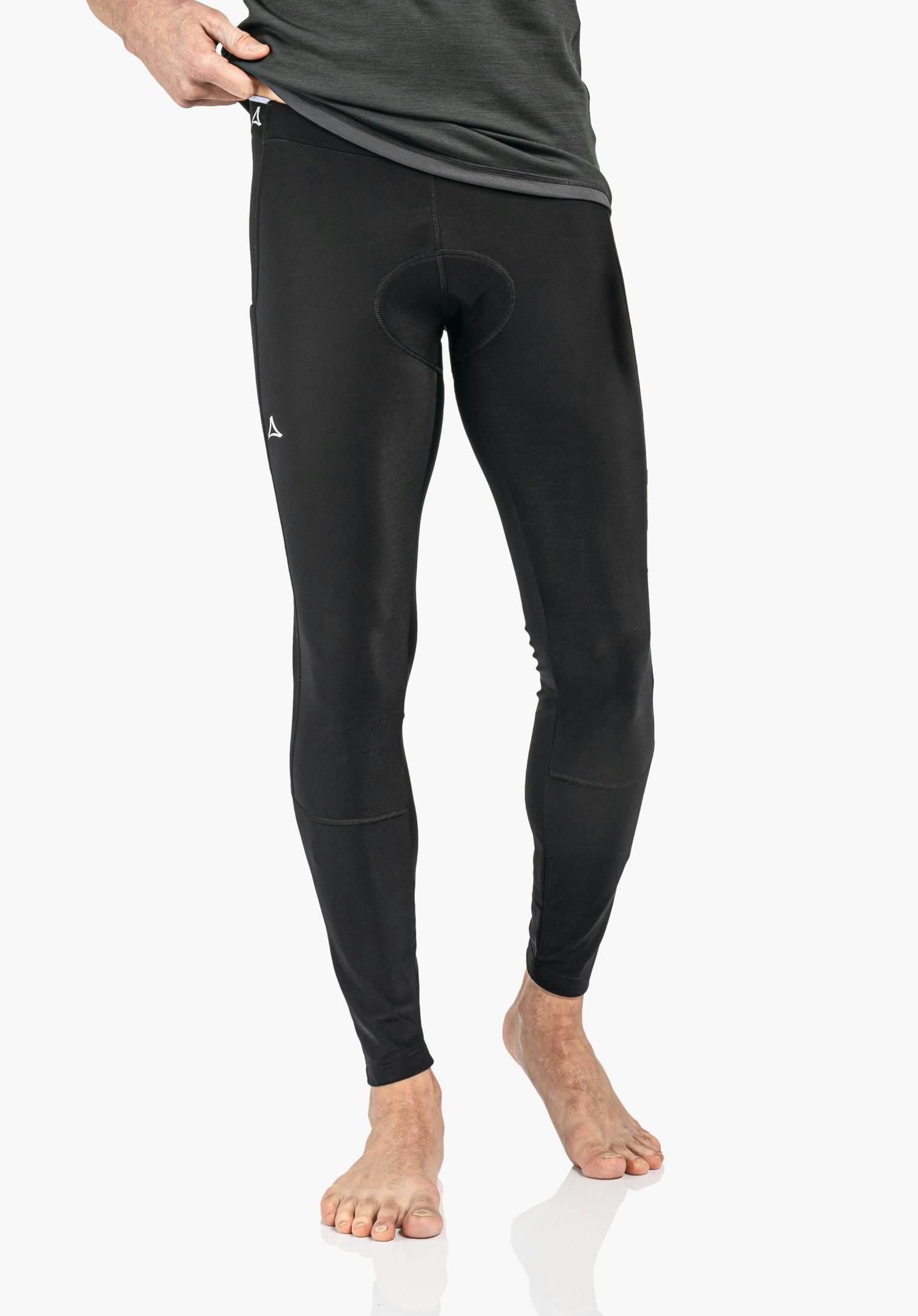 Skin Pants Warm 4h M Eng anliegende schwarze Leggings, von einer Hand am Bund hochgezogen, Schöffel-Logo weiß an Bund und Oberschenkel, Modell steht barfuß auf weißem Hintergrund.