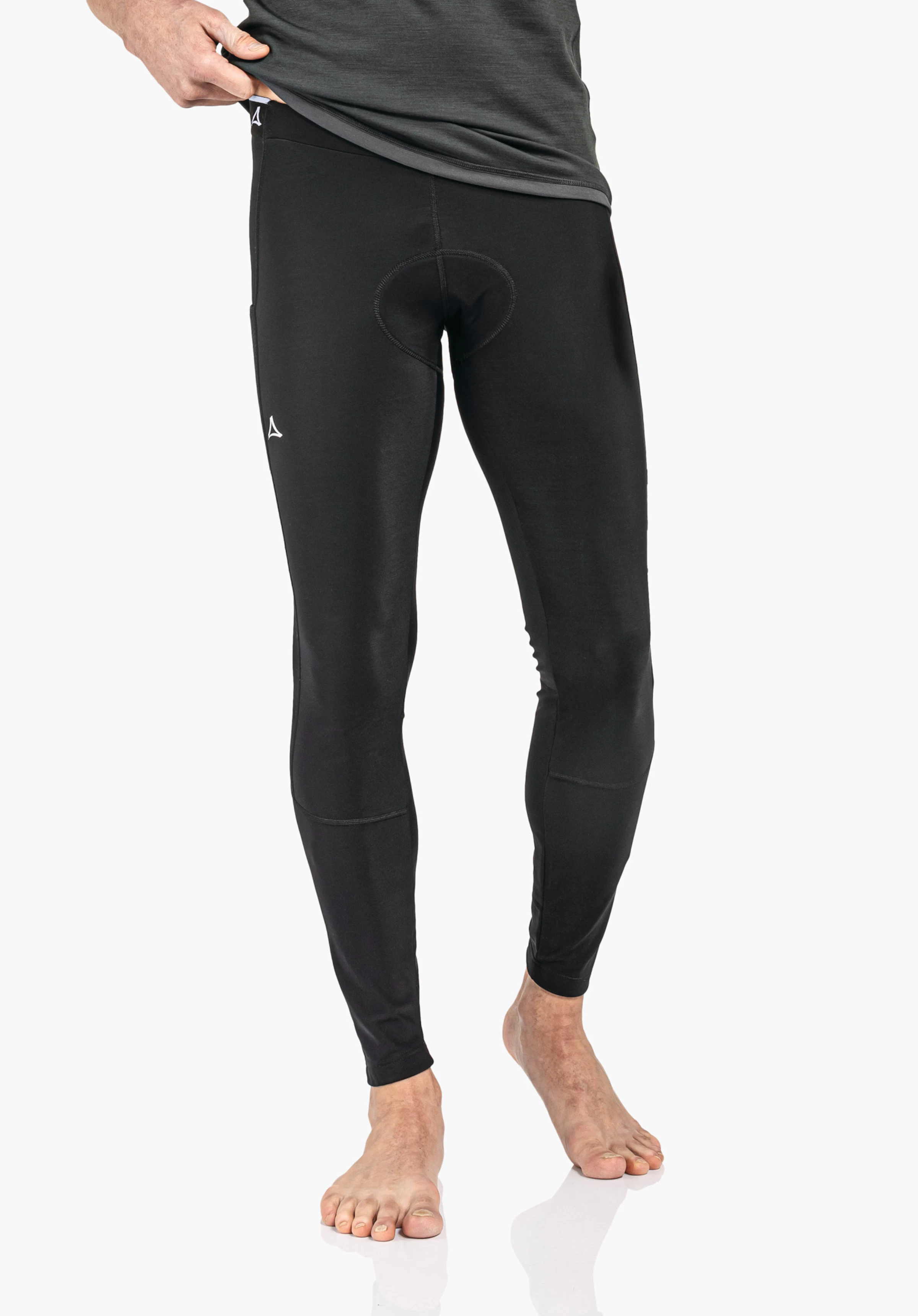 Skin Pants Warm 4h M Eng anliegende schwarze Leggings, von einer Hand am Bund hochgezogen, Schöffel-Logo weiß an Bund und Oberschenkel, Modell steht barfuß auf weißem Hintergrund.
