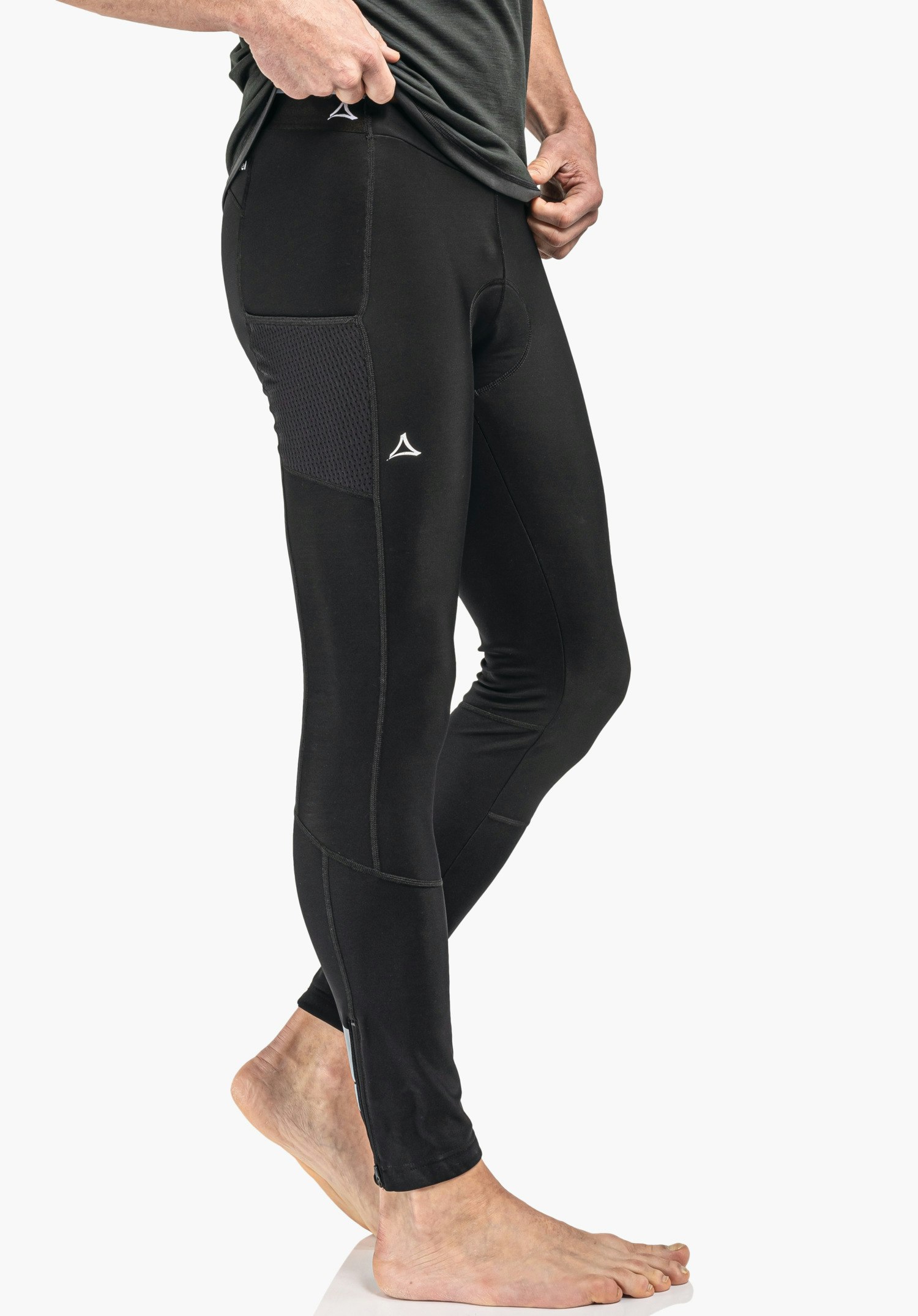 Skin Pants Warm 4h M Eng anliegende schwarze Leggings, Person hebt Oberteil an; seitliche Tasche und Reißverschluss am Knöchel, Schöffel-Logo auf Oberschenkel, barfüßig stehend vor weißem Hintergrund.