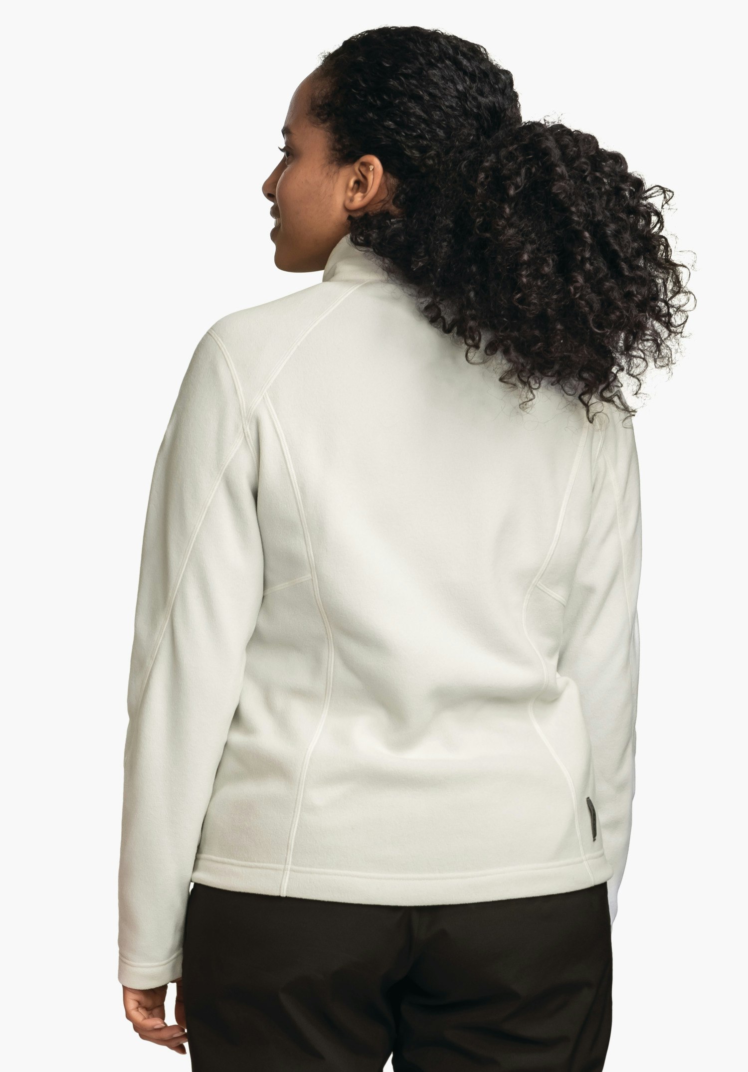 Fleece Jacket Leona3 lichtwitte fleecejas, gedragen door een persoon met zichtbare rug; persoon kijkt zijwaarts over de schouder, zwart krullend haar, donkerbruine broek, lichte neutrale achtergrond.