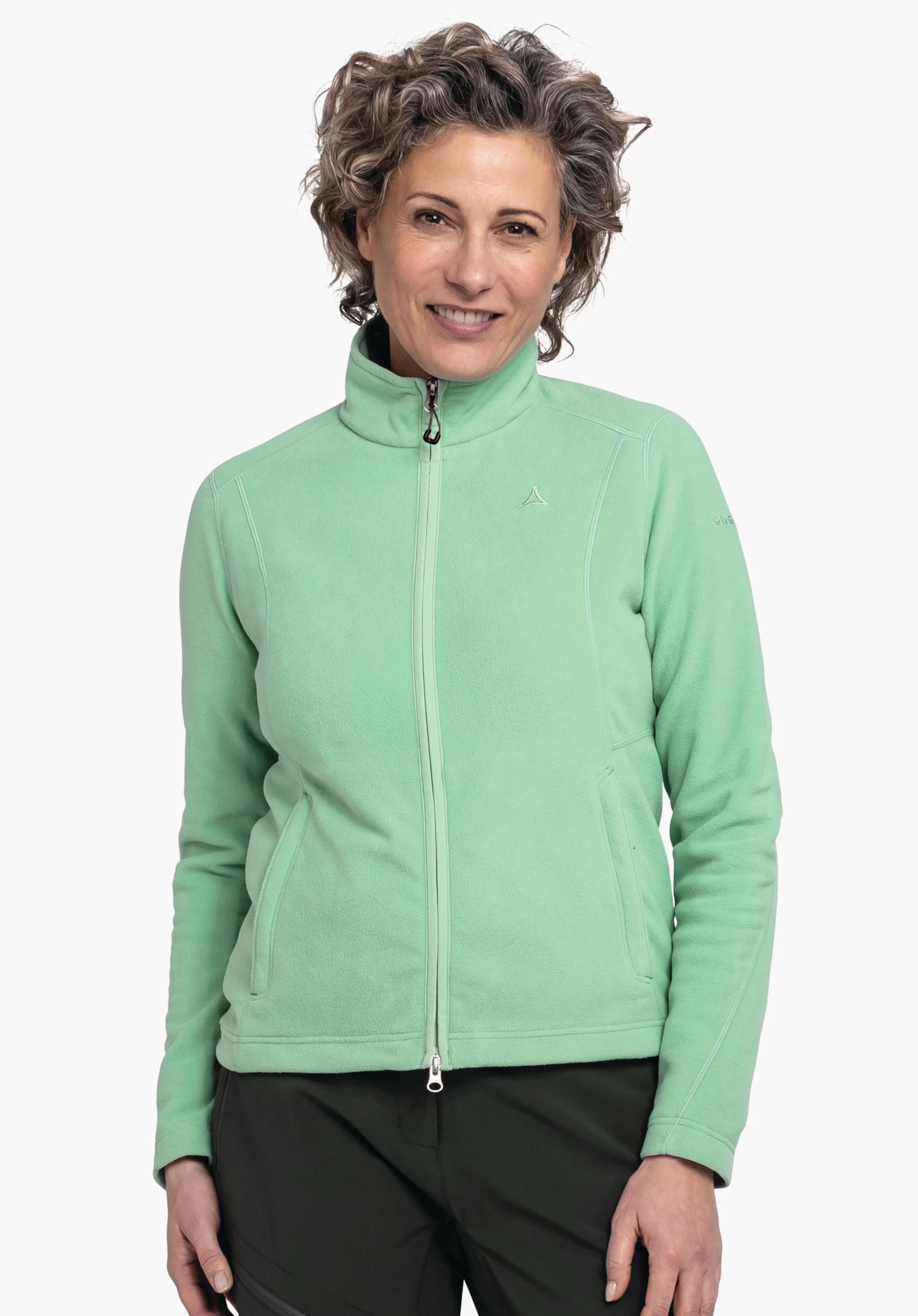 Fleece Jacket Leona3 Veste polaire vert clair avec zip et logo Schöffel sur la poitrine; associée à un pantalon vert foncé; modèle debout devant un fond blanc, bras détendus le long du corps.