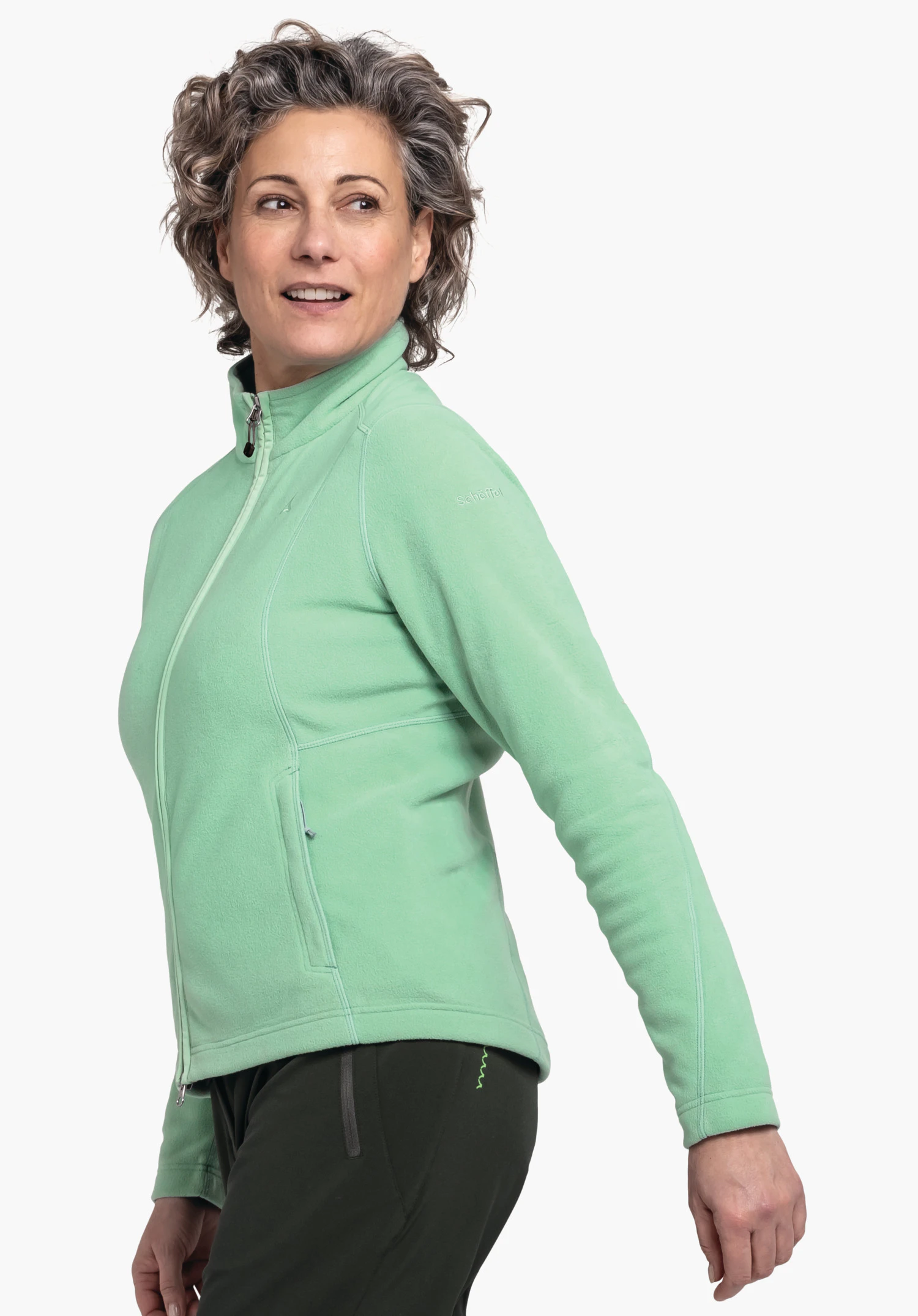 Fleece Jacket Leona3 veste polaire: femme tend le bras sur le côté, porte une veste vert clair cintrée avec fermeture éclair et un pantalon vert foncé devant fond blanc. Texte sur l’épaule: "Schöffel".