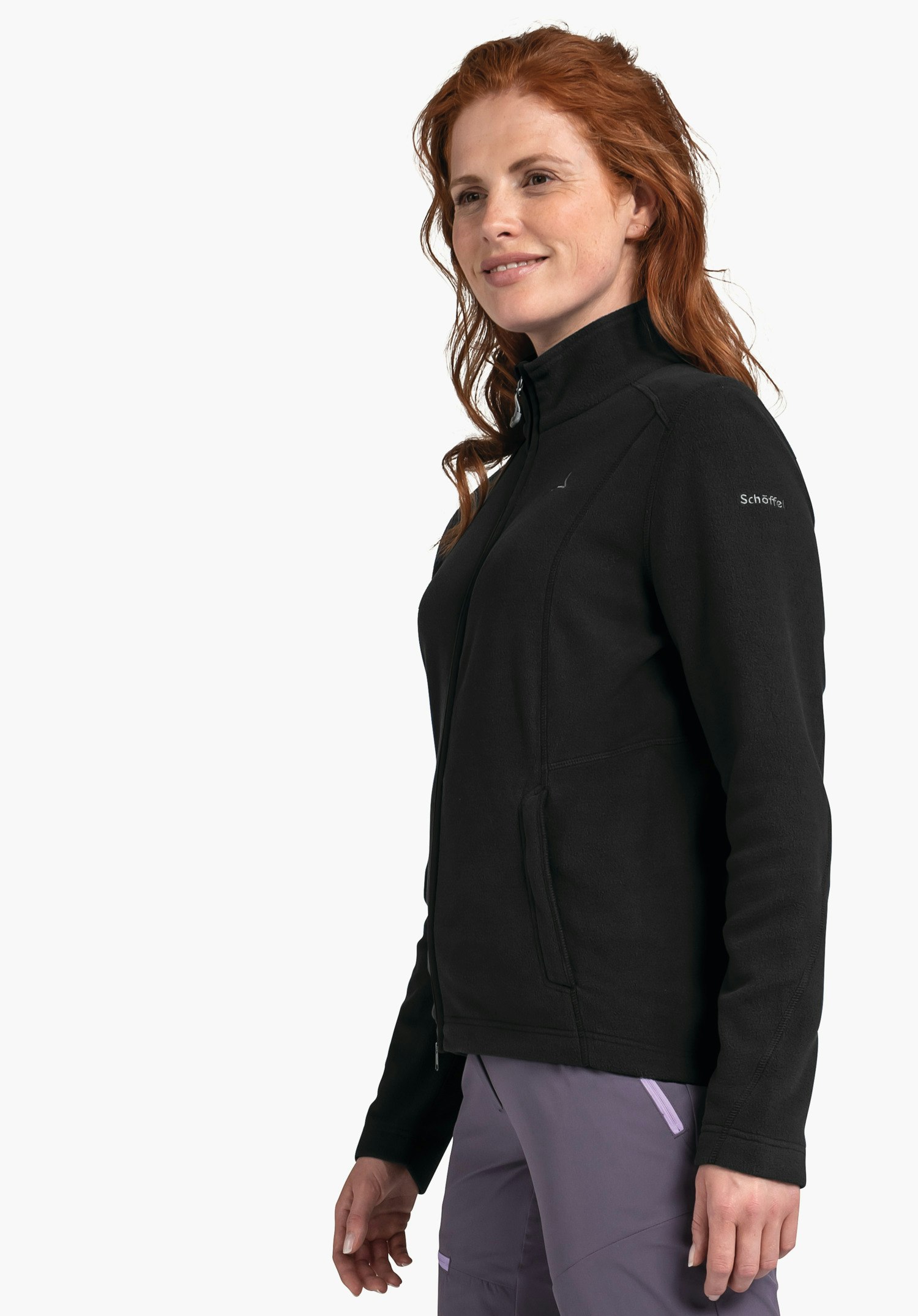 Fleece Jacket Leona3 veste polaire noire portée par une femme aux cheveux rouges; pose latérale devant fond blanc. Inscription sur la manche: "Schöffel". Pantalon violet.