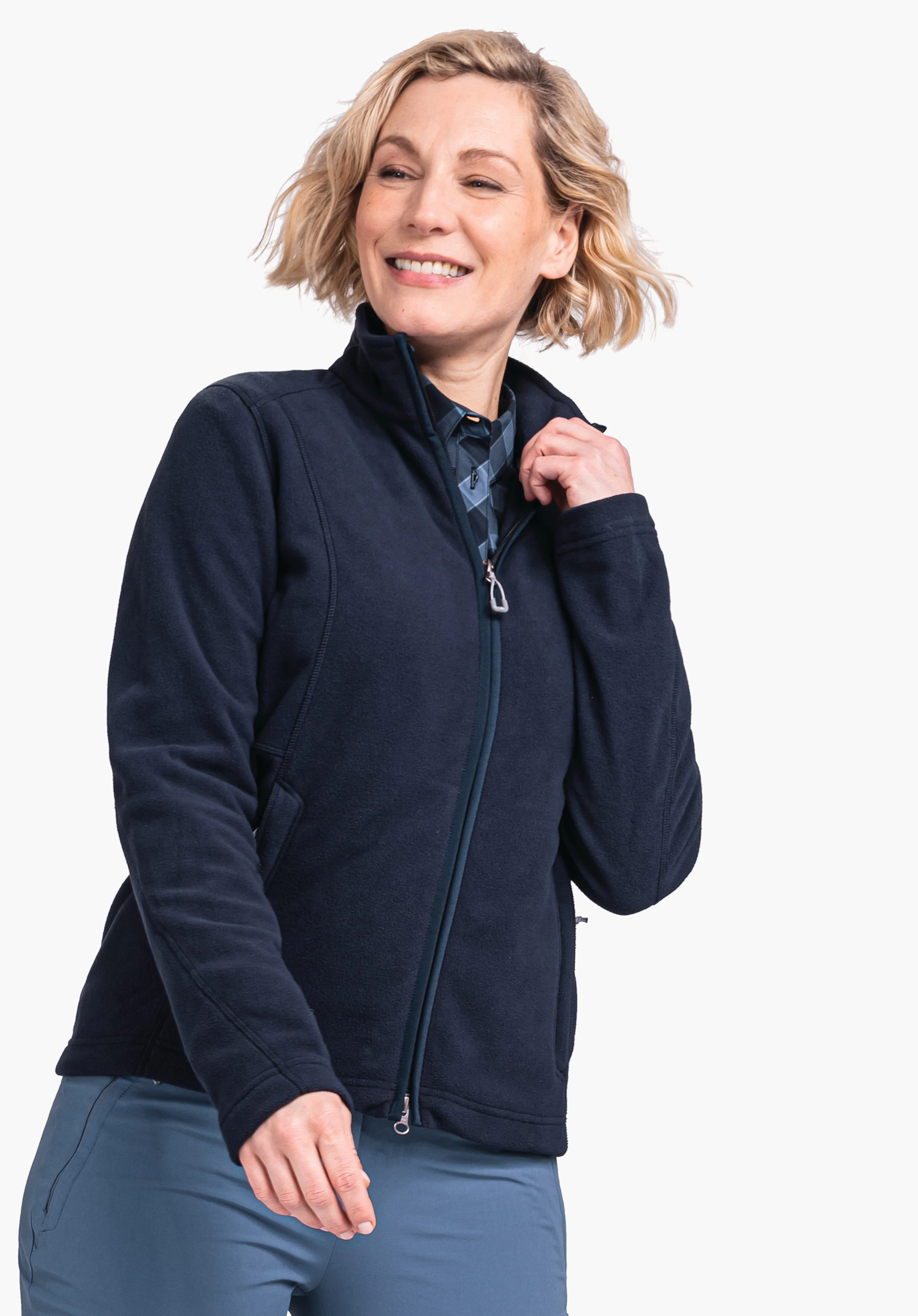 Fleece Jacket Leona3 donkerblauwe fleecejas; een vrouw trekt glimlachend de ritssluiting omhoog; zij draagt een lichtblauwe broek voor een lichte, effen achtergrond.
