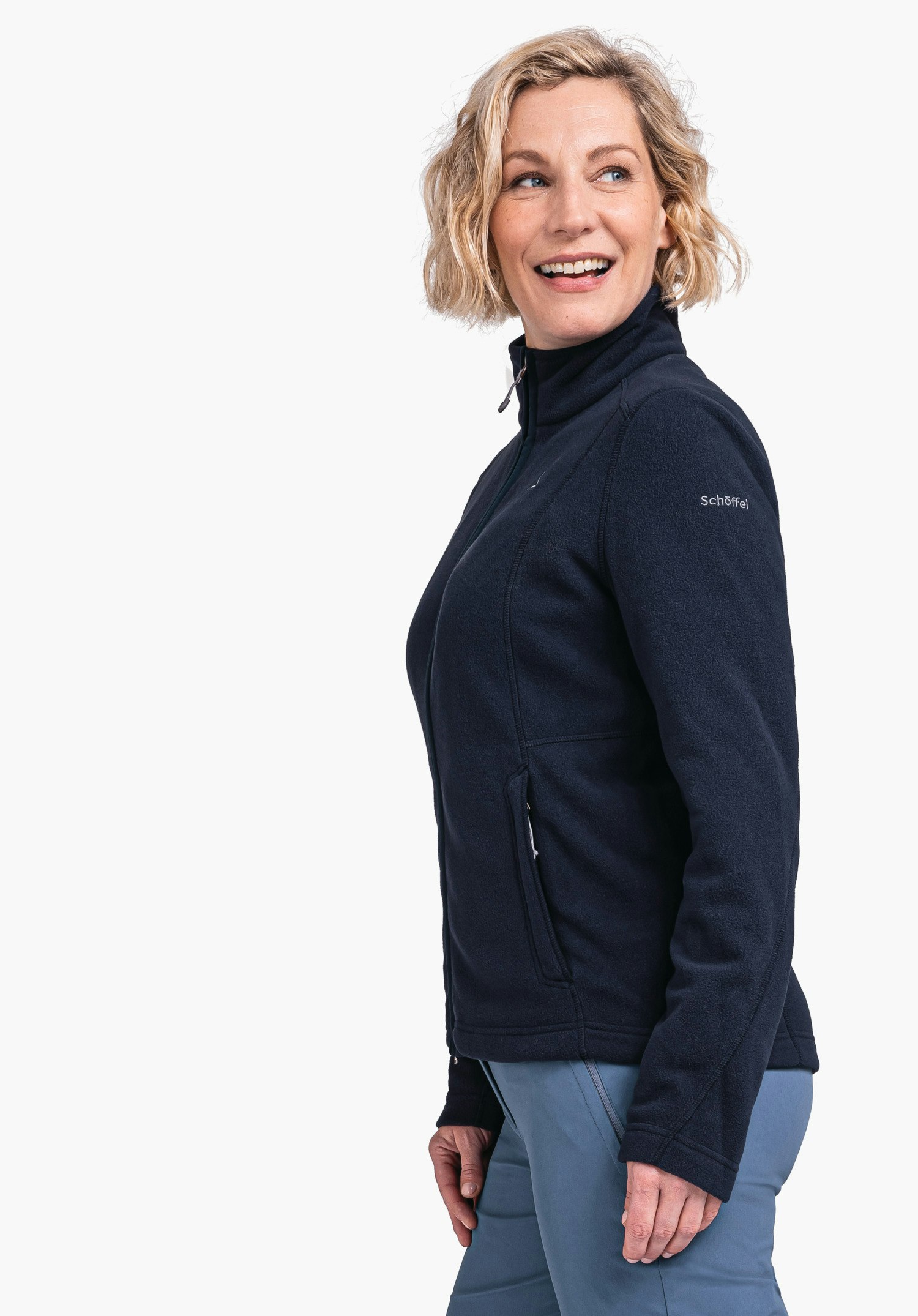 Fleece Jacket Leona3 woman wears a dark blue fleece jacket, smiles and looks side-on; standing in front of a white background. Light blue trousers. Sleeve text: "Schöffel".