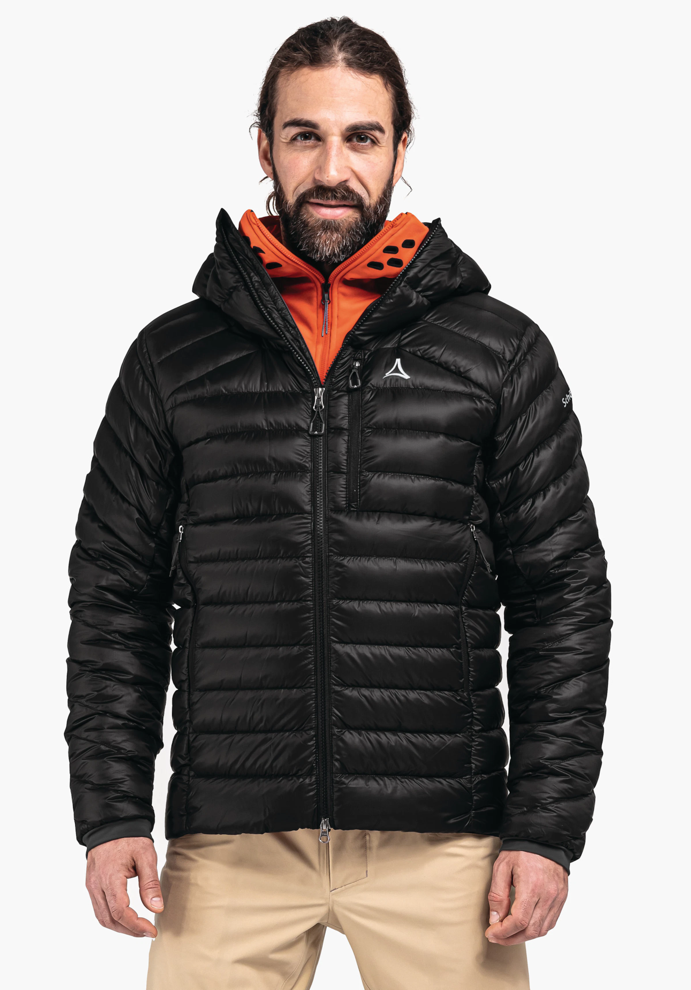 Down Jacket Silvretta M Mann mit schwarzer Steppjacke (Schöffel-Logo auf Brust) steht frontal und lächelt; Kapuzeninnenseite hell orange, trägt beige Hosen vor weißem Hintergrund. Text: "Schöffel".