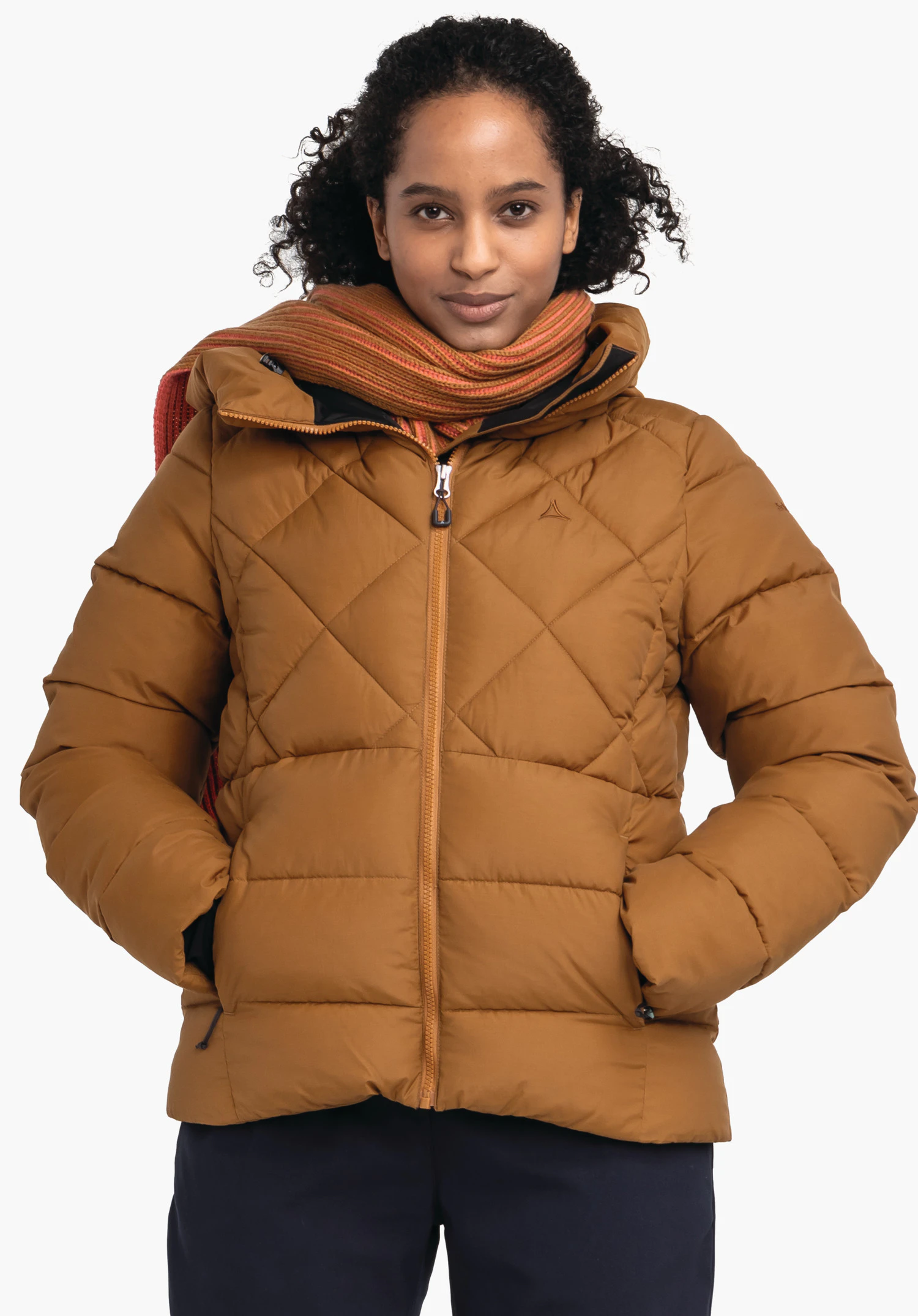 Ins Jacket Boston L dunkelbraune Steppjacke mit Schöffel-Logo, getragen von einer Frau; sie hat die Hände in den Taschen und trägt einen orange und rot gestreiften Schal, vor weißem Hintergrund.