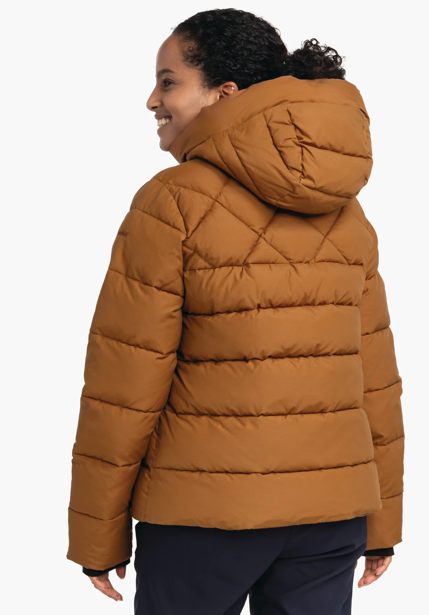 Ins Jacket Boston L donkeroranje gewatteerde jas met capuchon, van achteren gedragen door een glimlachende persoon die zijwaarts kijkt; lichte studioachtergrond, donkerblauwe broek zichtbaar.