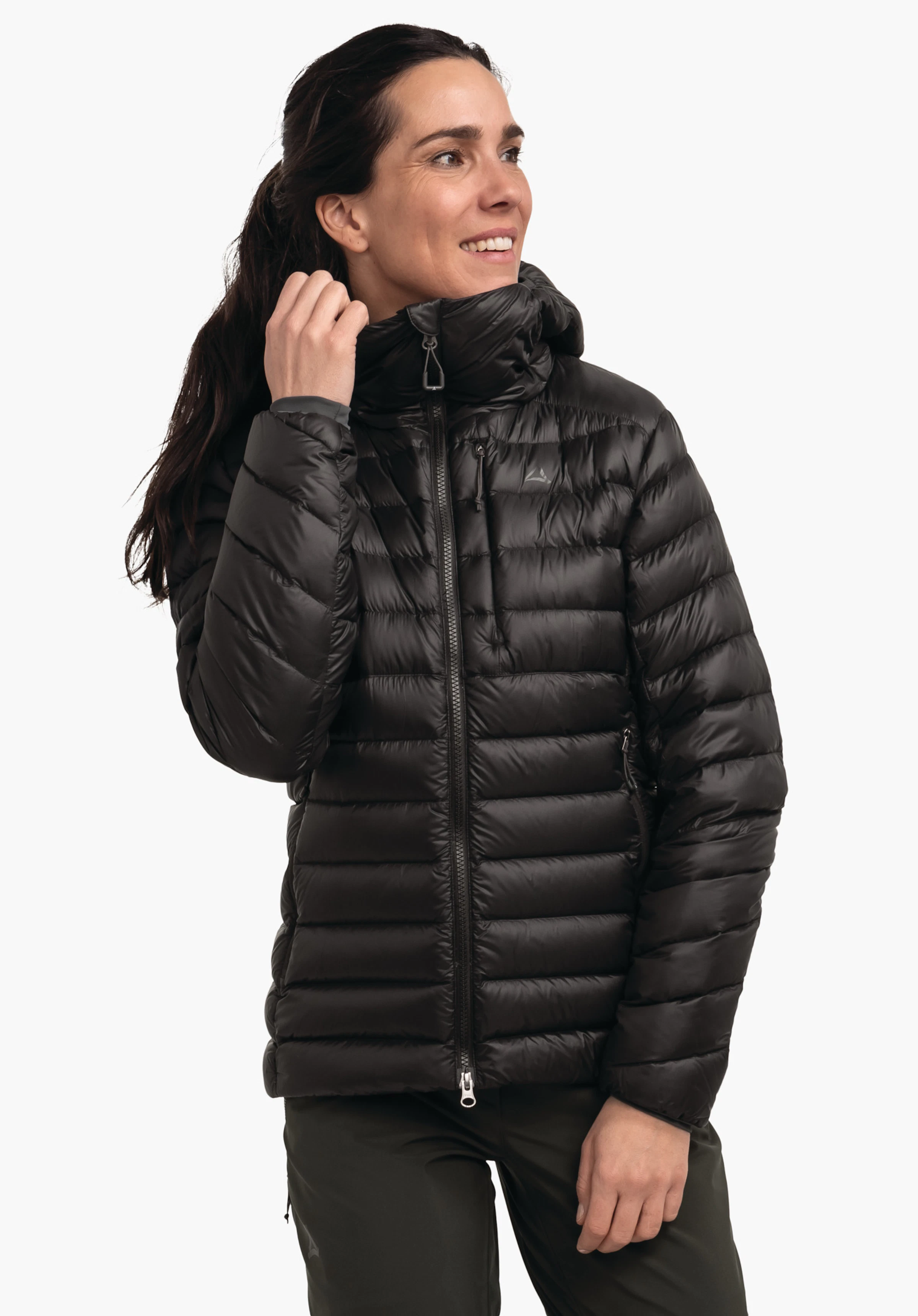 Down Jacket Silvretta L Schwarze gesteppte Daunenjacke, getragen von einer Frau, die den hohen Kragen fasst; Reißverschluss, Kapuze, horizontale Steppnähte; heller Hintergrund; Schöffel-Logo auf der Brust.