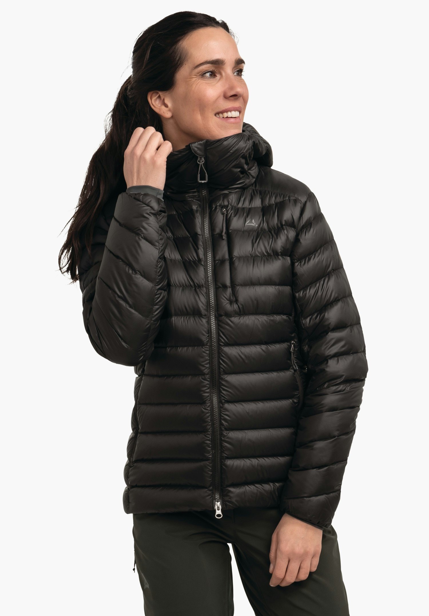 Down Jacket Silvretta L doudoune noire matelassée, portée par une femme tenant le col montant; fermeture éclair, capuche, matelassage horizontal; fond clair; logo Schöffel sur la poitrine.