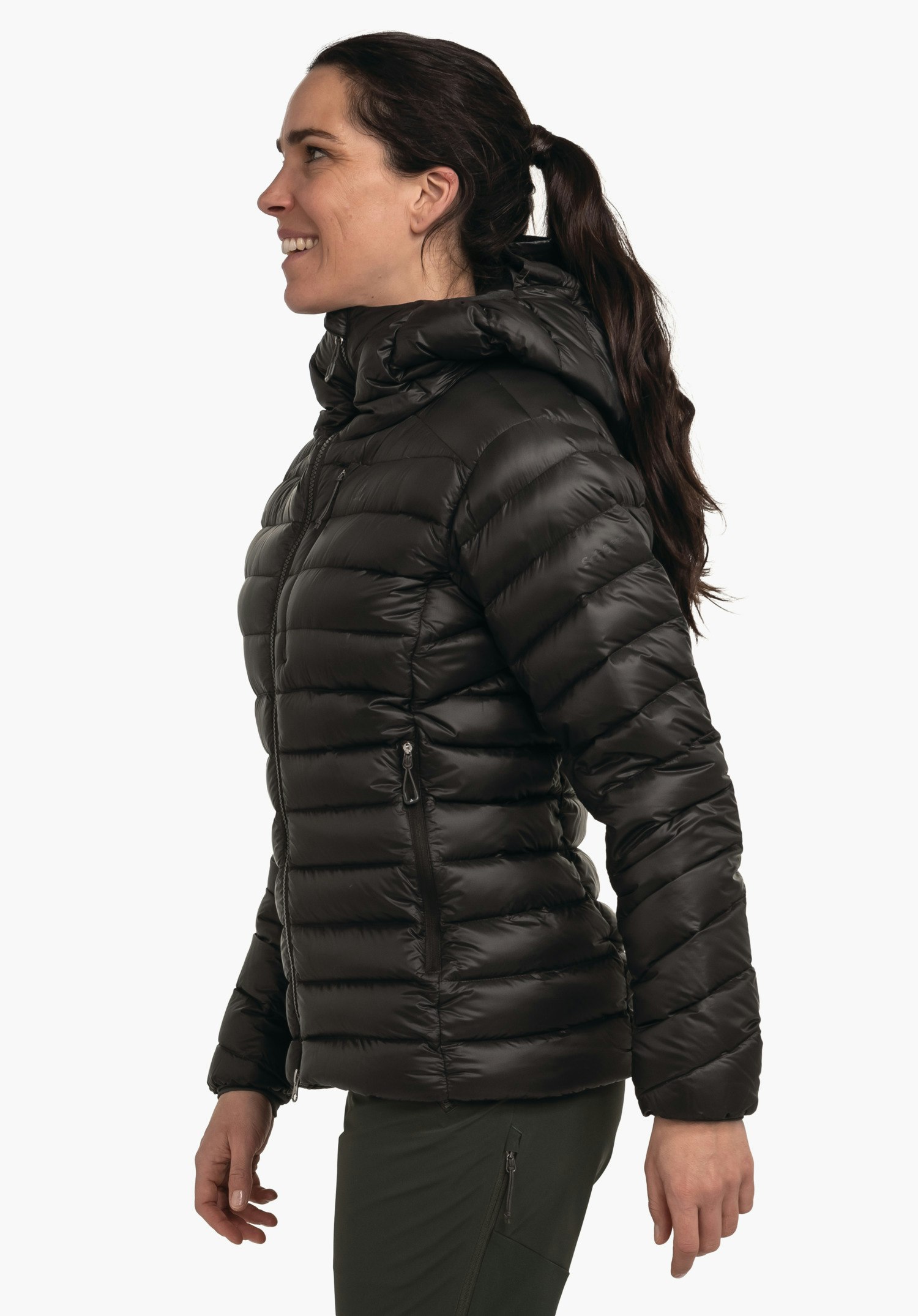 Down Jacket Silvretta L doudoune noire portée par une femme debout en profil et souriante; veste avec capuche et poches zippées; fond blanc clair, pantalon vert foncé.