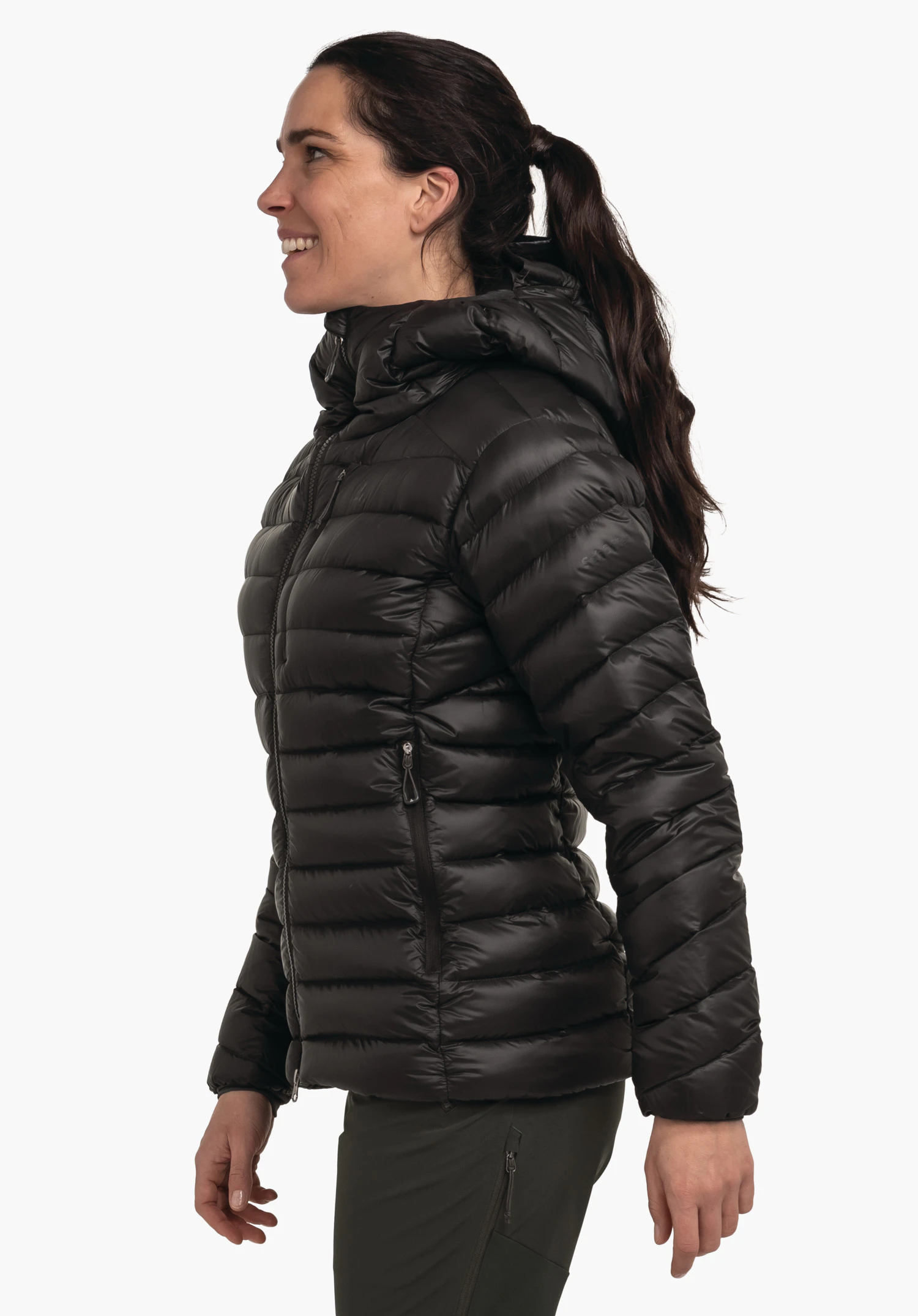 Down Jacket Silvretta L Schwarze Daunenjacke, von einer Frau getragen, die im Profil lächelnd steht; Jacke mit Kapuze und Reißverschlusstaschen; Hintergrund weiß hell, Hose grün dunkel.