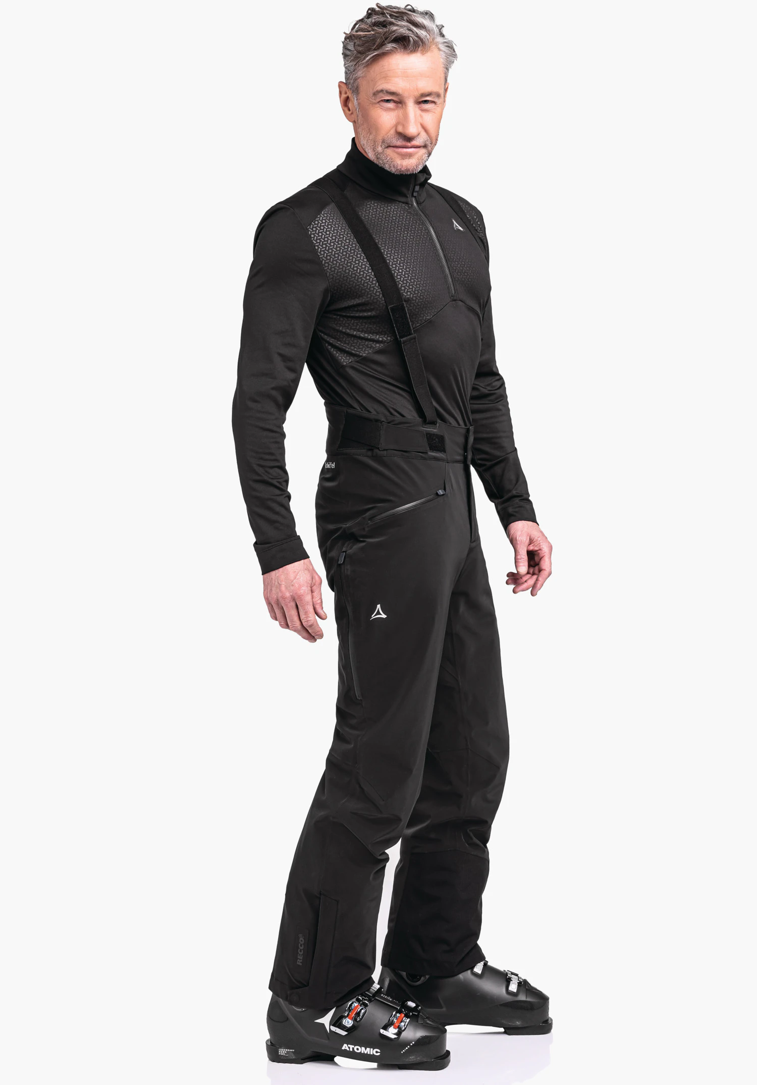 Ski Pants Trevalli M man wears a black ski jacket and trousers with Schöffel logo, stands side-on with a relaxed stance and looks ahead; white background. Text in image: ATOMIC; RECCO.