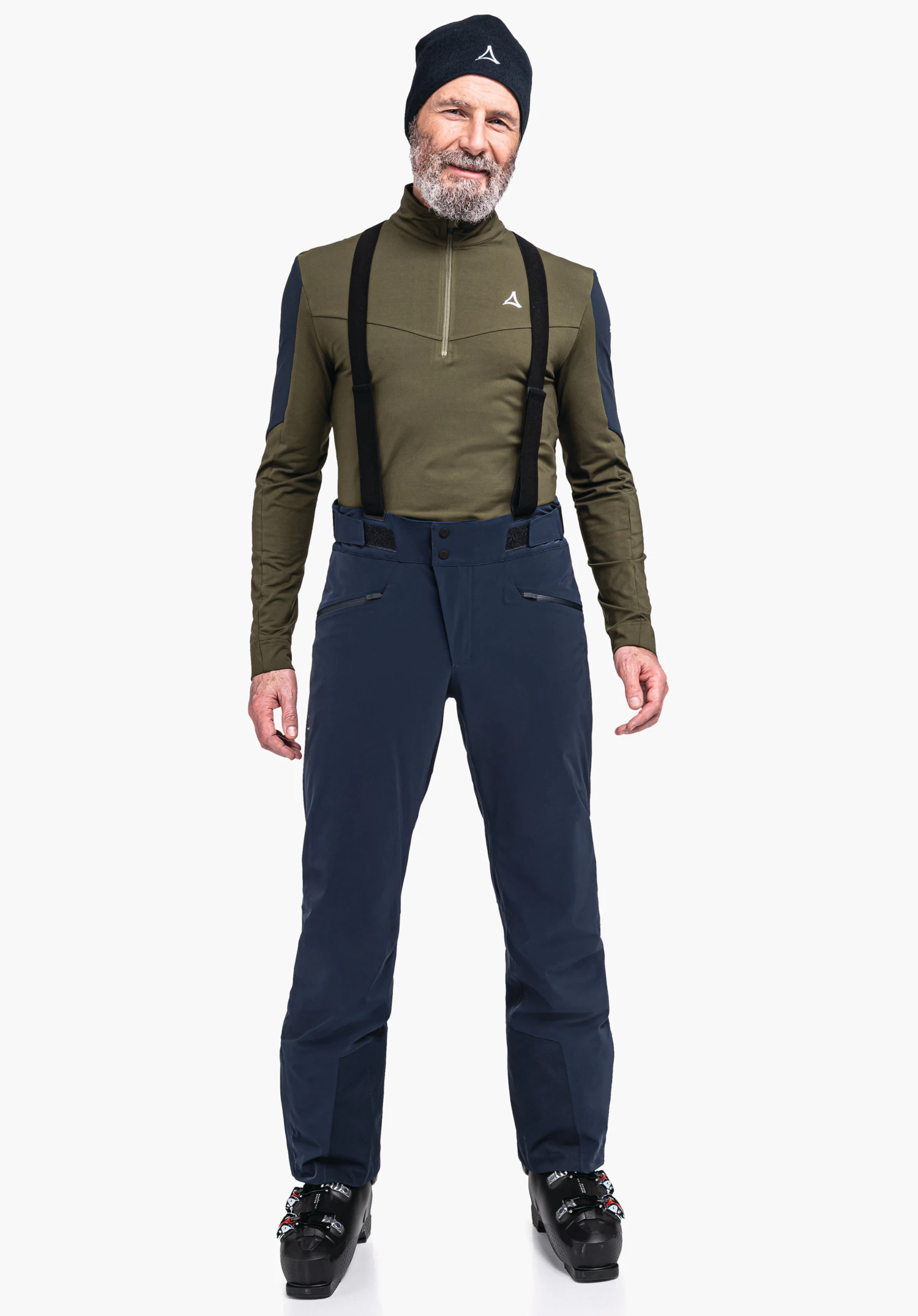Ski Pants Trevalli M homme portant un haut vert foncé avec logo Schöffel sur la poitrine et un bonnet noir avec logo Schöffel, bretelles noires; debout en pantalon de ski bleu foncé et chaussures de ski noires devant un fond clair.