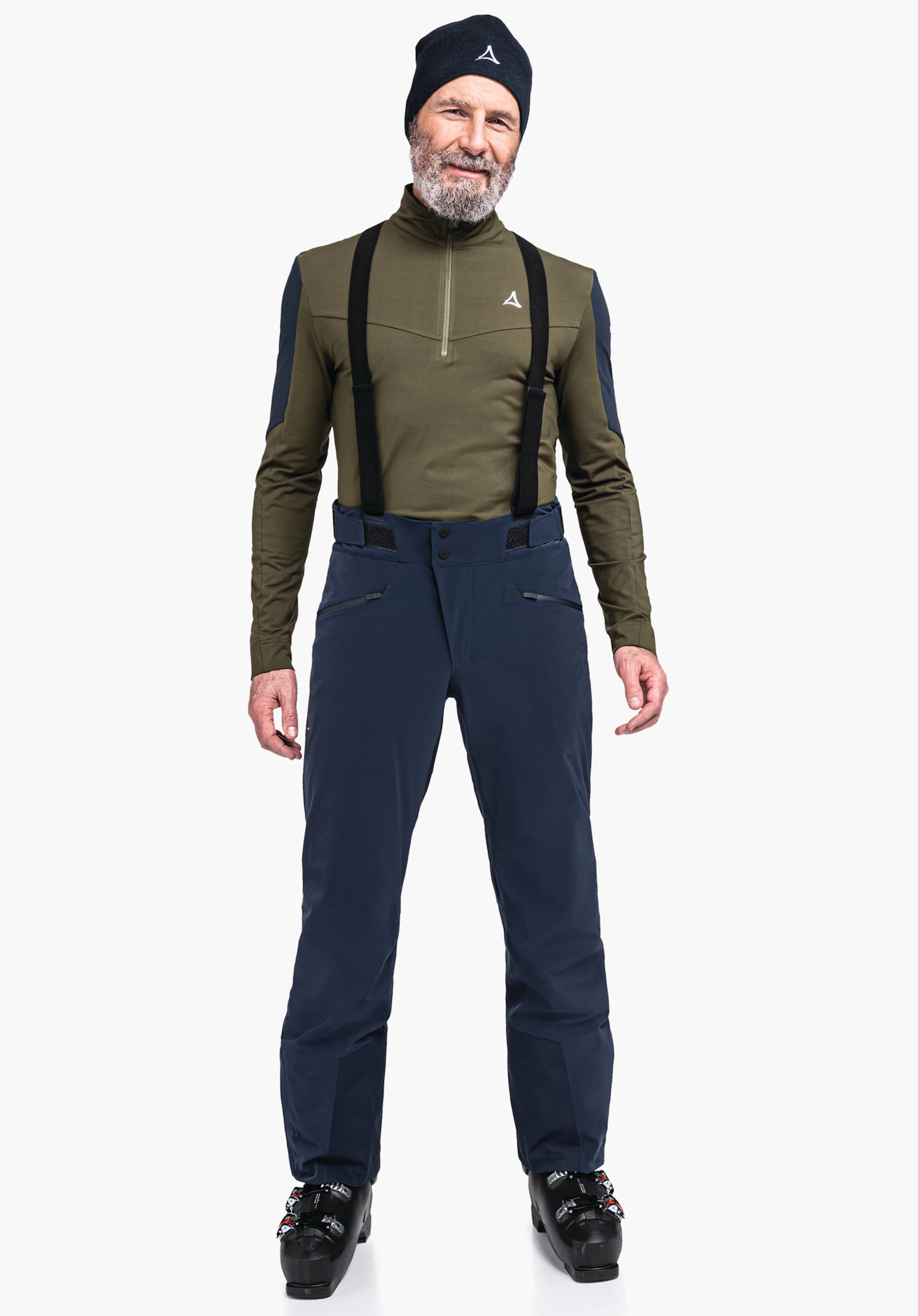 Ski Pants Trevalli M man wearing a dark green top with Schöffel logo on the chest and a black beanie with Schöffel logo, black braces; standing in dark blue ski trousers and black ski boots against a light background.