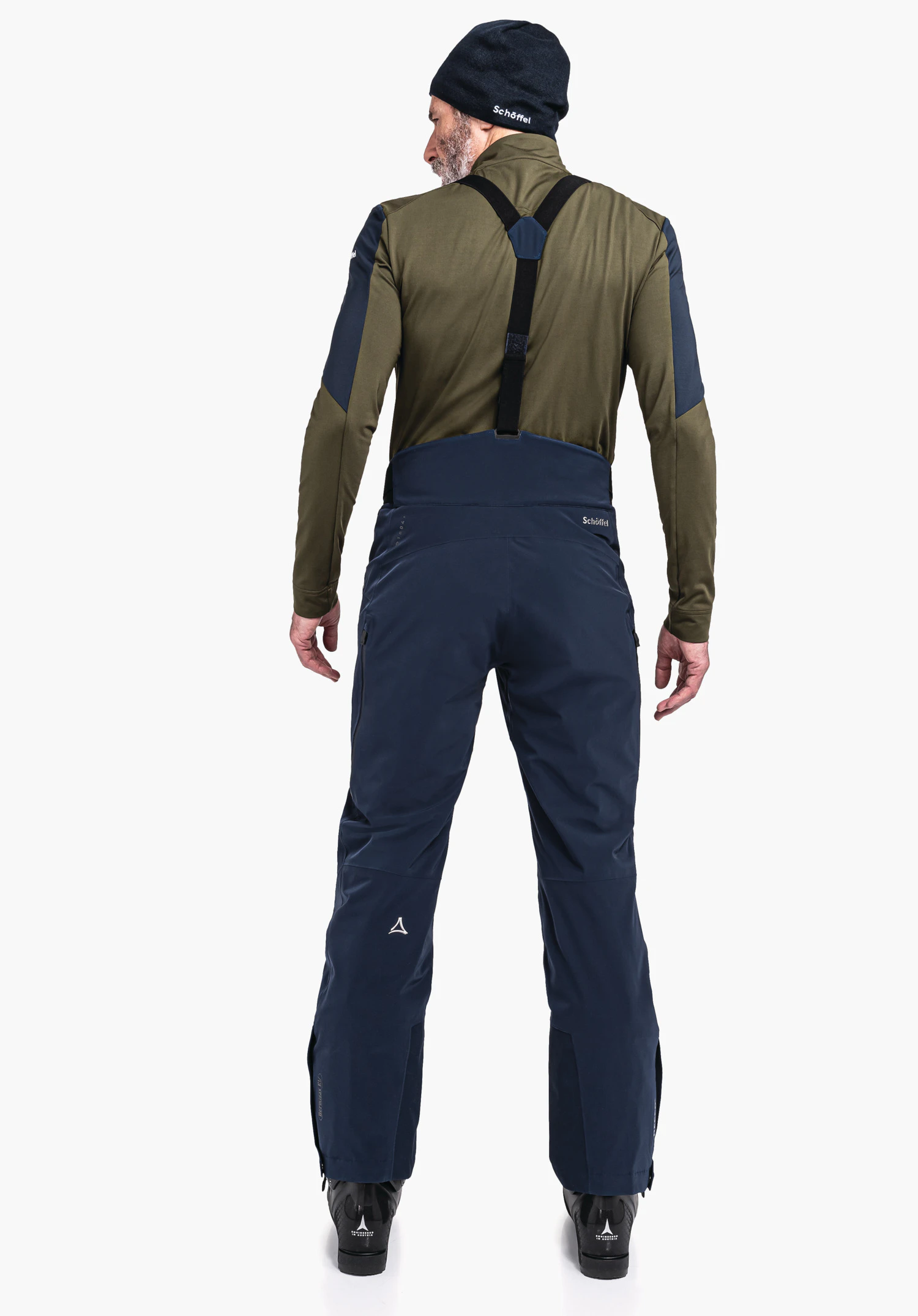 Ski Pants Trevalli M homme debout dos à la caméra, portant un pantalon de ski bleu foncé avec bretelles noires, un haut manches longues vert foncé et un bonnet bleu foncé devant un fond clair. Texte: "Schöffel". Logo Schöffel visible.