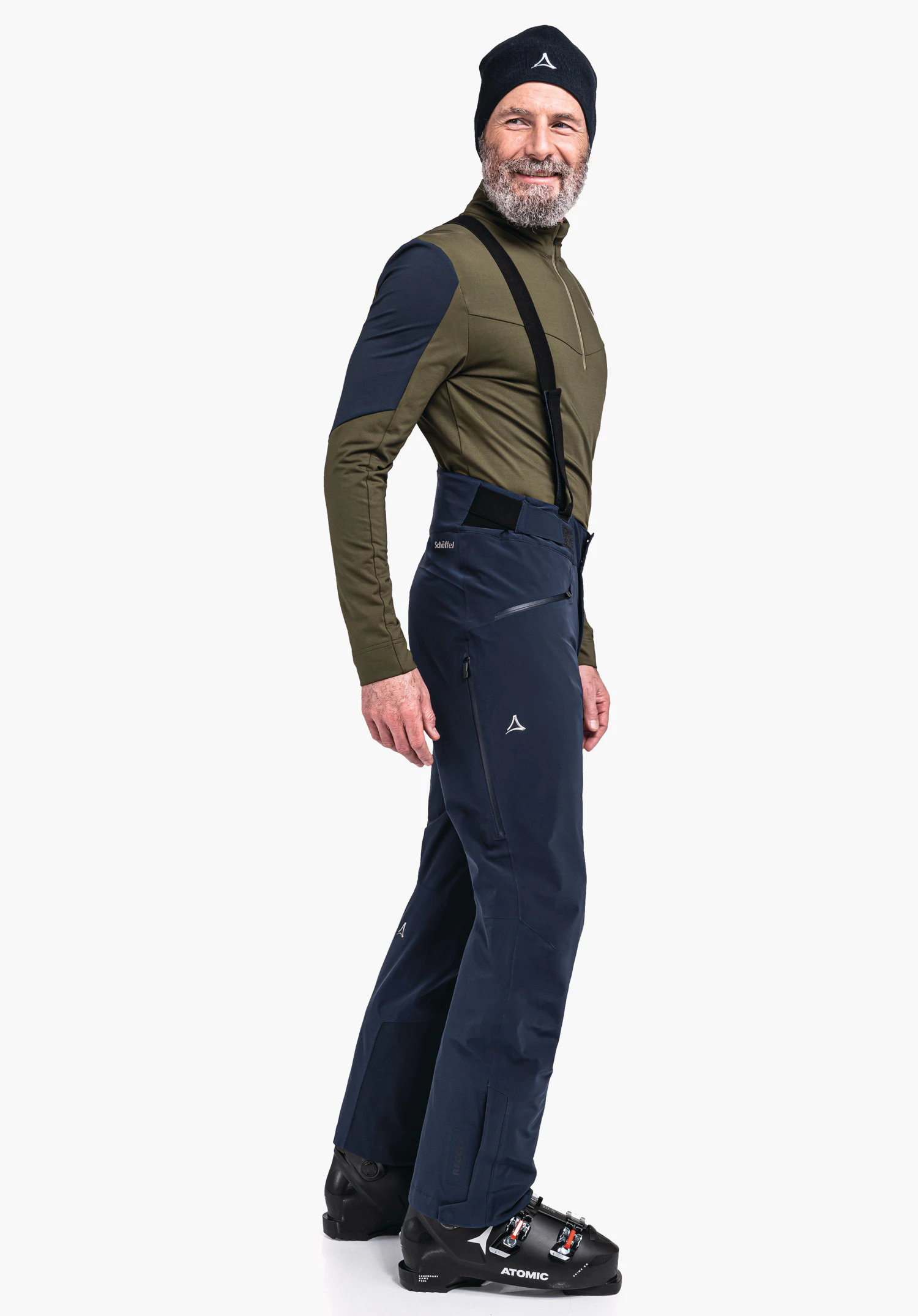 Ski Pants Trevalli M homme portant un haut manches longues vert foncé avec empiècements bleu foncé, pantalon de ski bleu foncé avec logo Schöffel, bonnet noir et chaussures de ski noires; debout de profil et souriant devant fond clair. Texte: "Schöffel", "Atomic".
