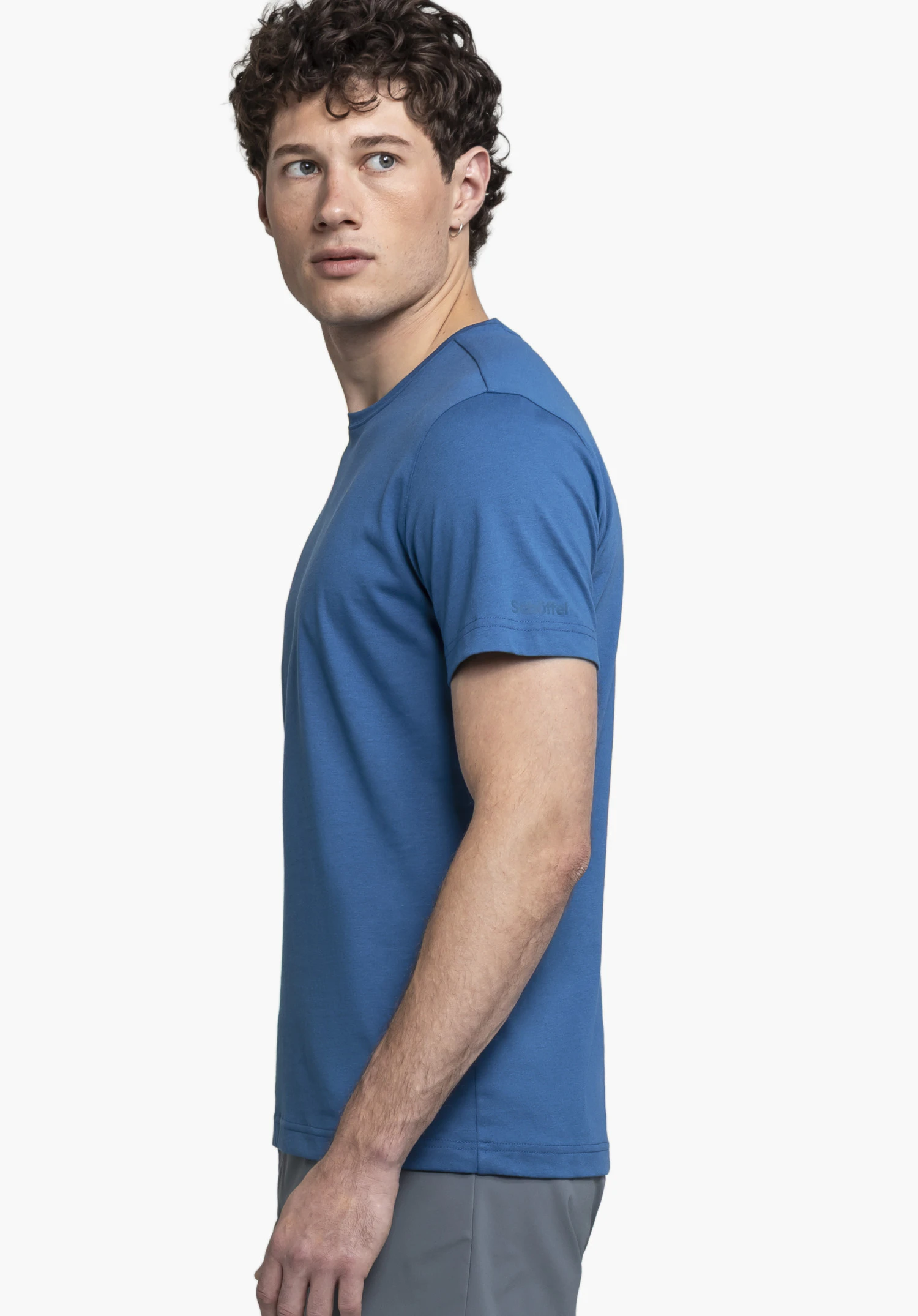 T Shirt Buchberg M en bleu. Le mannequin se tient de profil devant un fond neutre et regarde directement vers l'appareil photo. Le design épuré est subtilement mis en valeur par le logo Schöffel ton sur ton sur la manche.