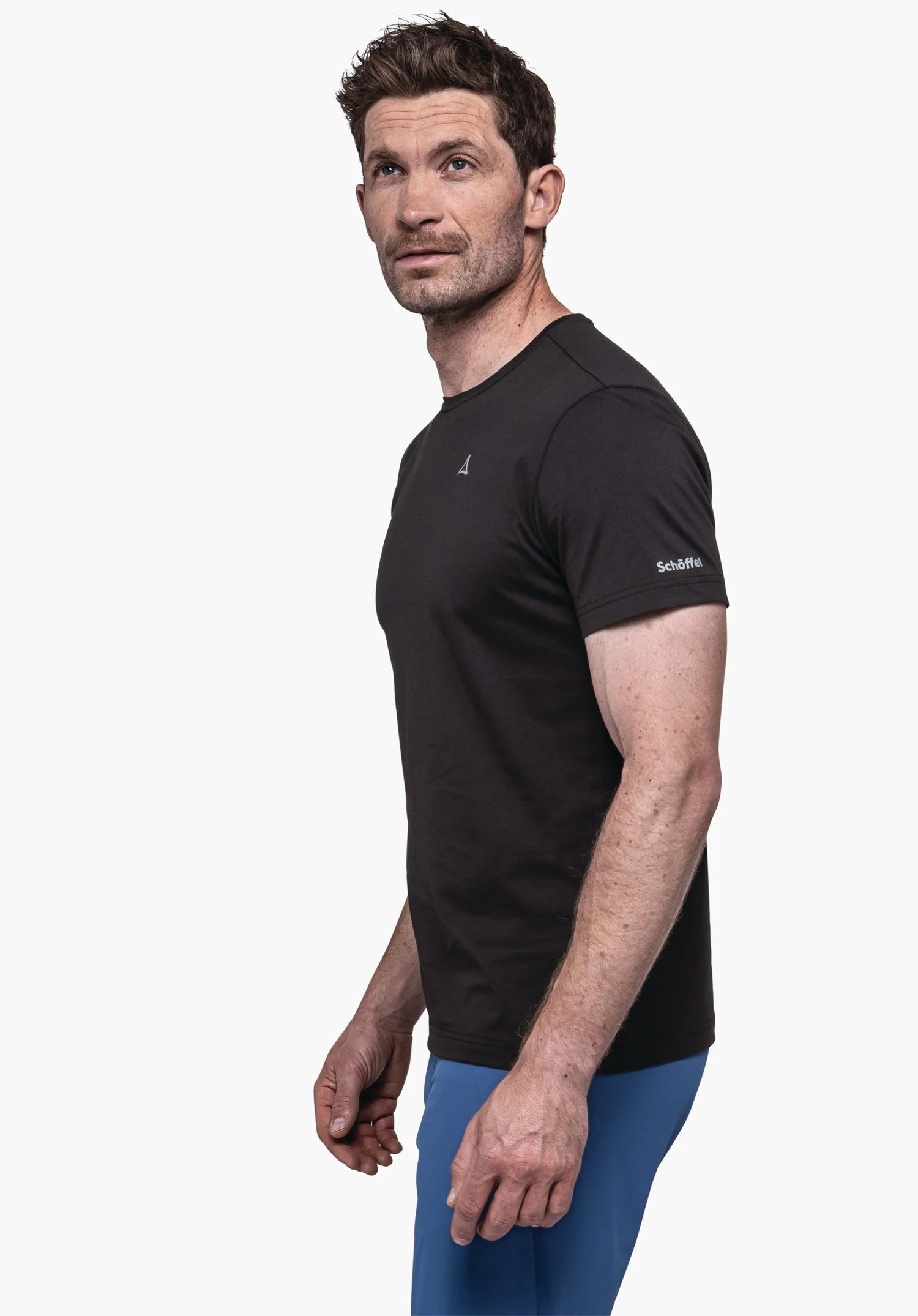 T Shirt Hohberg M in het zwart. Het mannelijke model staat in zijaanzicht voor een neutrale achtergrond en kijkt opzij. Het zwarte T-shirt heeft korte mouwen en is voorzien van een subtiel Schöffel-logo op de mouw.