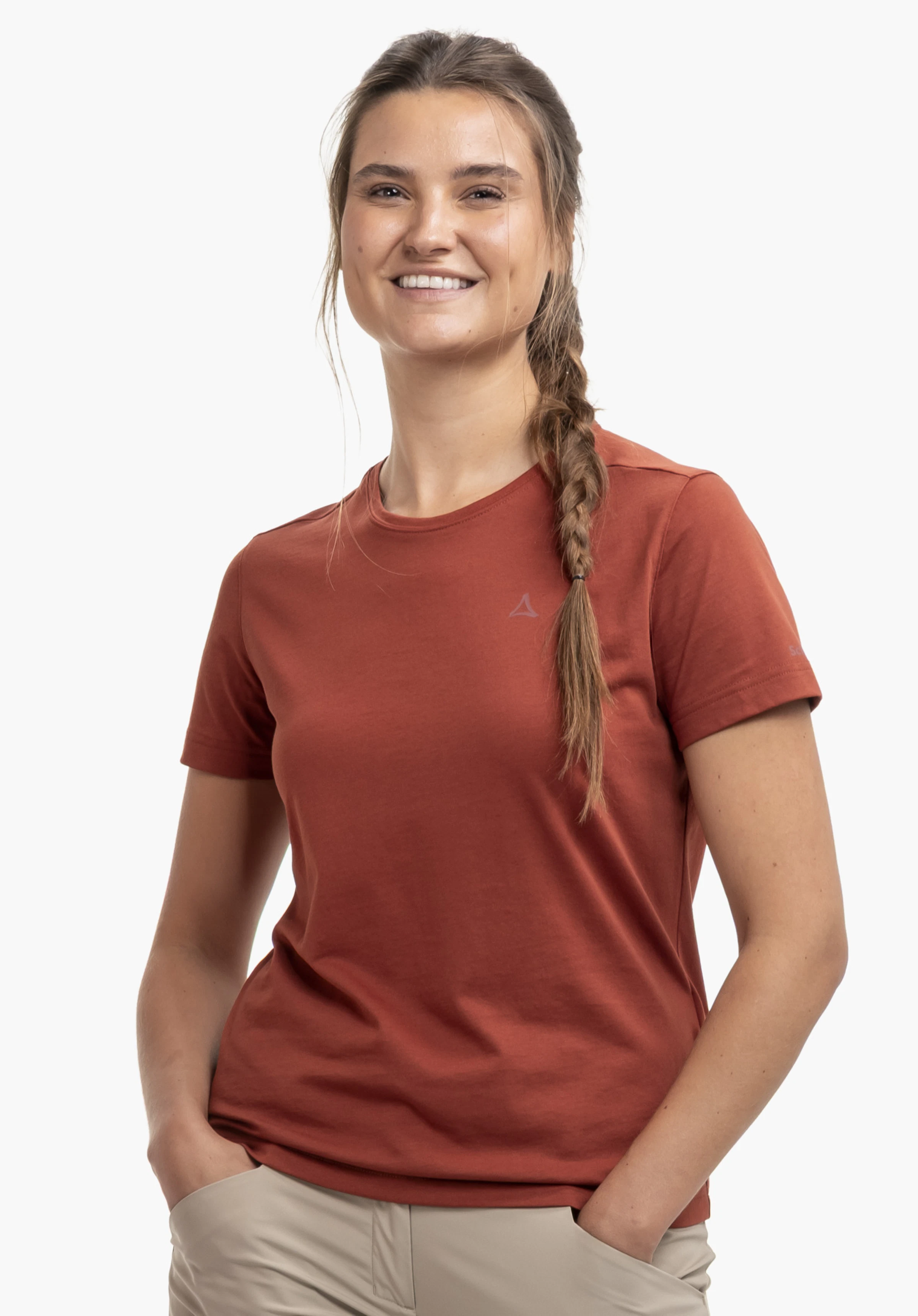 T Shirt Hohberg L en rouge, porté par un mannequin souriant de face à la caméra. Elle a les mains glissées de manière décontractée dans les poches de son pantalon. Le logo Schöffel est bien visible sur le côté gauche de la poitrine. Photographié sur un fond neutre.