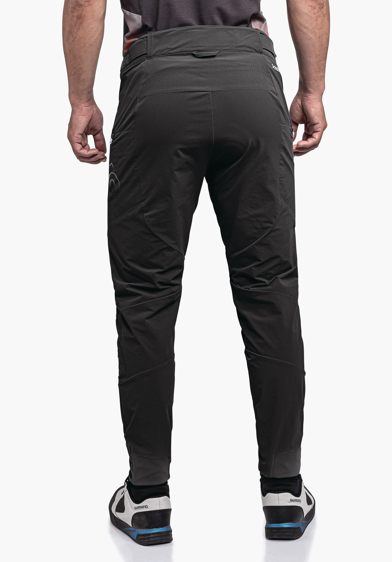 Pants Arosa M pantalon gris foncé, porté par une personne debout, vue arrière; coupe technique et ajustée avec poches latérales, chaussures blanches à détails noirs et bleus. Texte: "schöffel", "SHIMANO".