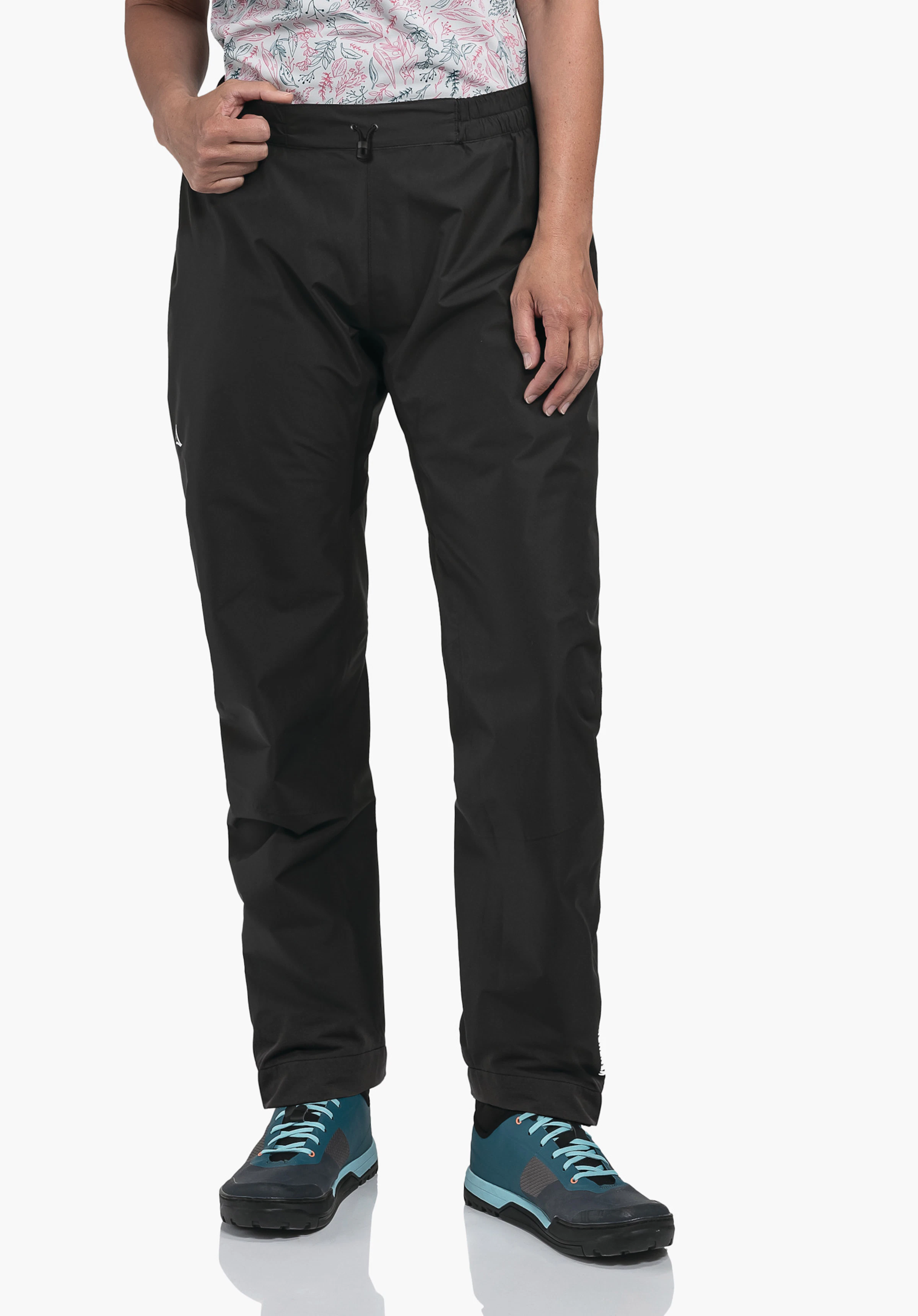 2.5L Pants Tarvis L very dark black trousers; person stands adjusting the waistband with the left hand; white shirt with light red floral pattern; dark blue shoes with light blue details; Schöffel logo on the left thigh.