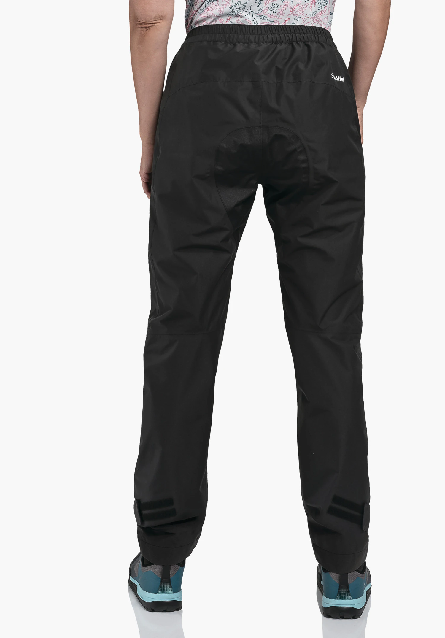 2.5L Pants Tarvis L black rain trousers worn from the back; standing with legs slightly apart, Velcro fastenings at the hems; neutral white background. Text: “Schöffel”.