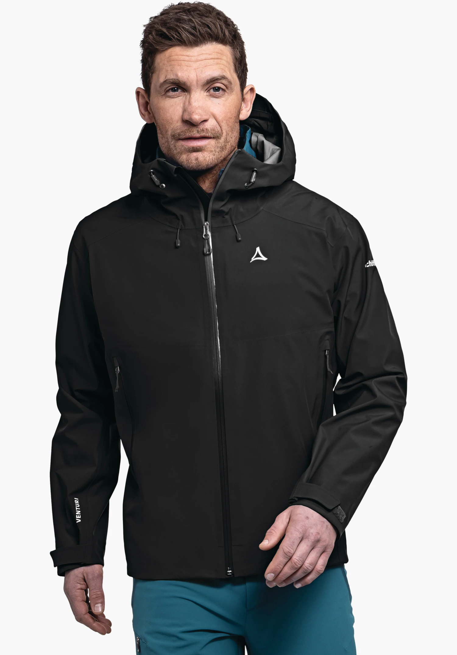 Jacket Style Cascata MNS Veste noire avec logo Schöffel sur la poitrine, portée par un homme; impression sur la manche: VENTURI; combinée avec un pantalon bleu clair devant un fond blanc.