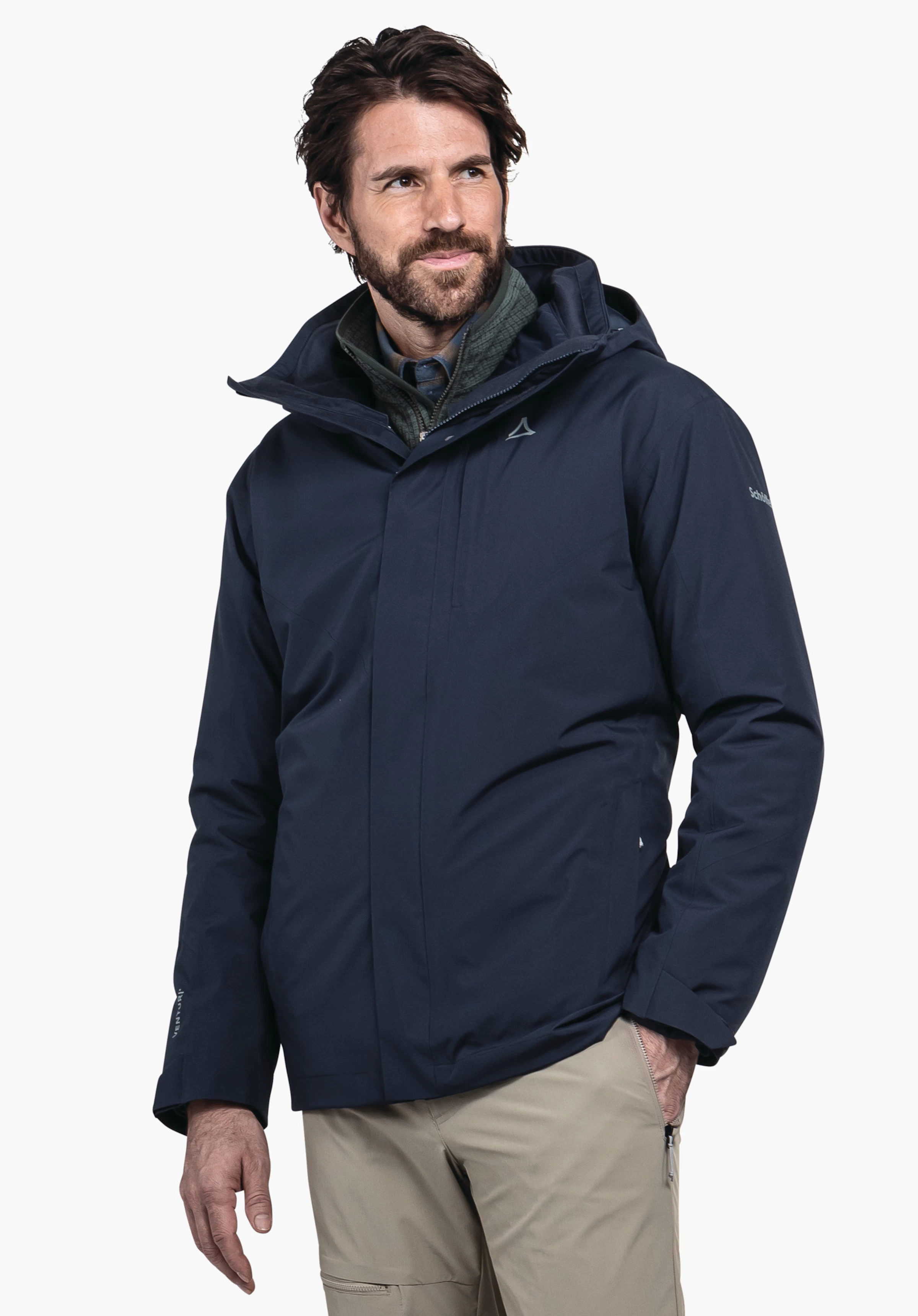 3in1 Jacket Style Tamina MNS veste outdoor bleu foncé, portée par un homme regardant vers la droite; logo Schöffel sur la poitrine, couche intérieure vert foncé, pantalon beige, fond blanc. Texte: «Schöffel».