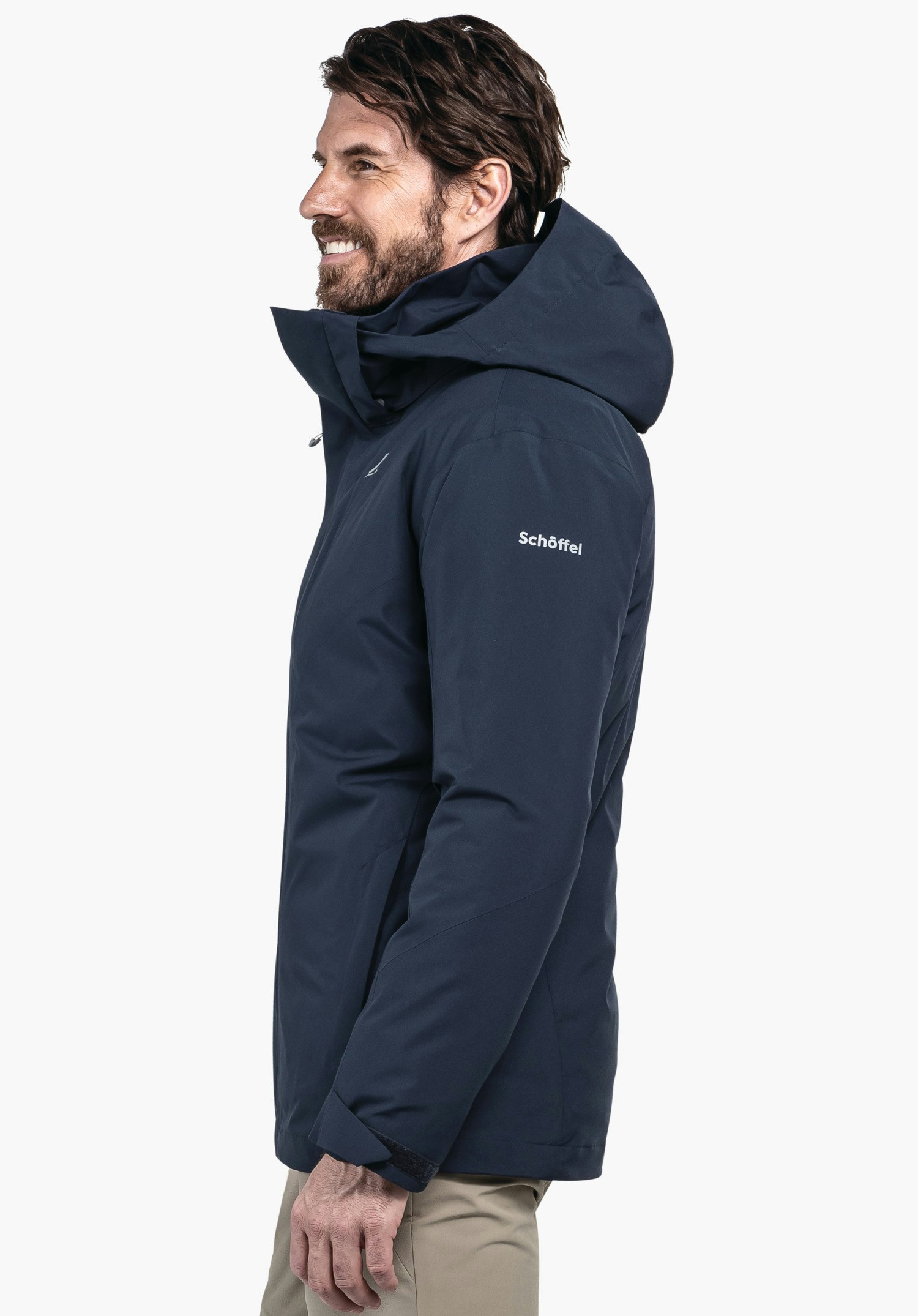 3in1 Jacket Style Tamina MNS Dunkelblaue Jacke, Mann trägt sie seitlich, lächelnd; Kapuze aufgesetzt; Kontext: neutraler heller Hintergrund. Text auf Kleidungsstück: "Schöffel". Schöffel-Logo sichtbar.
