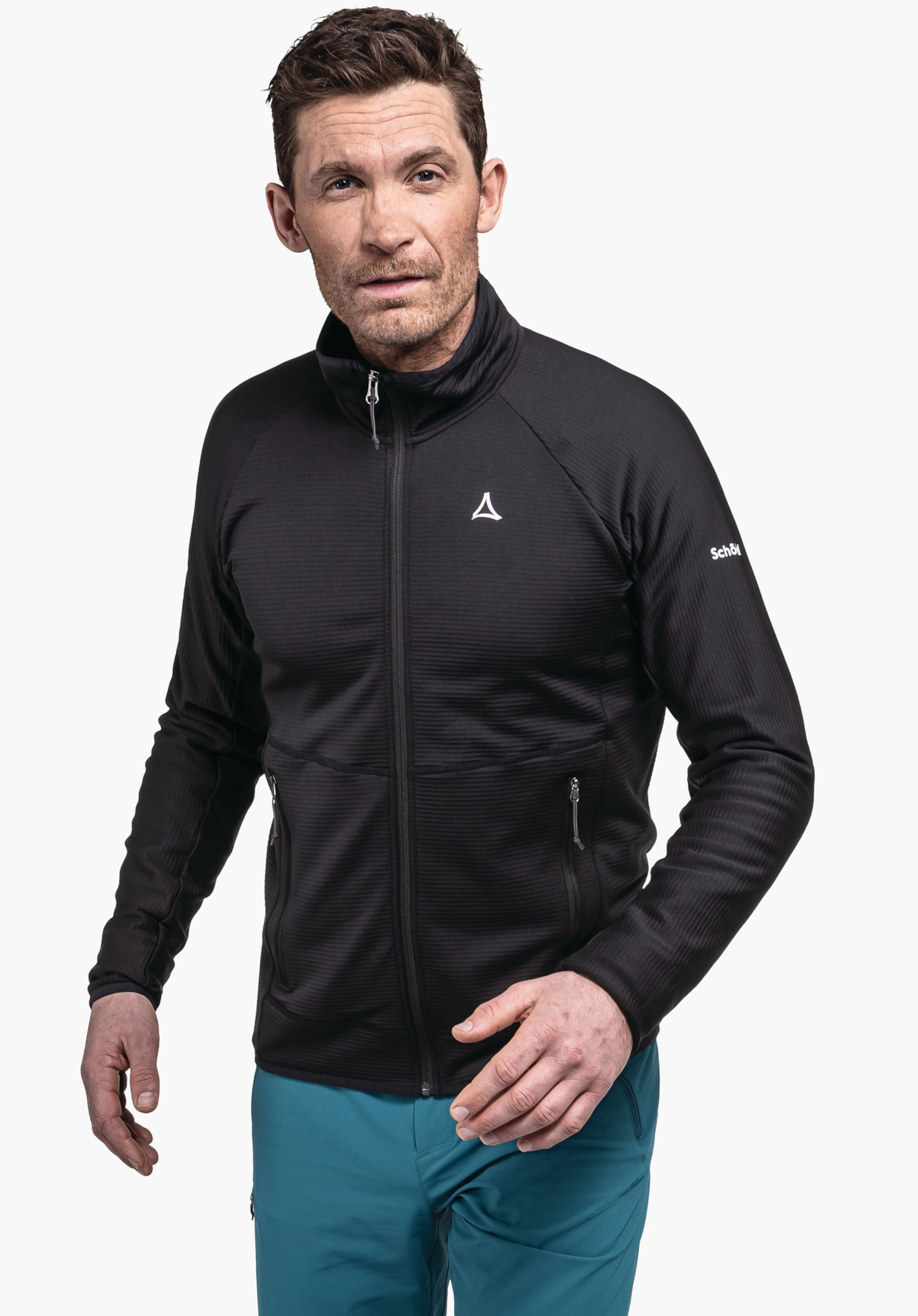 Fleece Jk Style Cascata MNS veste noire à col montant, portée par un homme; fermeture éclair fermée, logo Schöffel sur la poitrine; mains légèrement relevées; pantalon bleu clair; fond clair et neutre; texte sur la manche: «Schöffel».