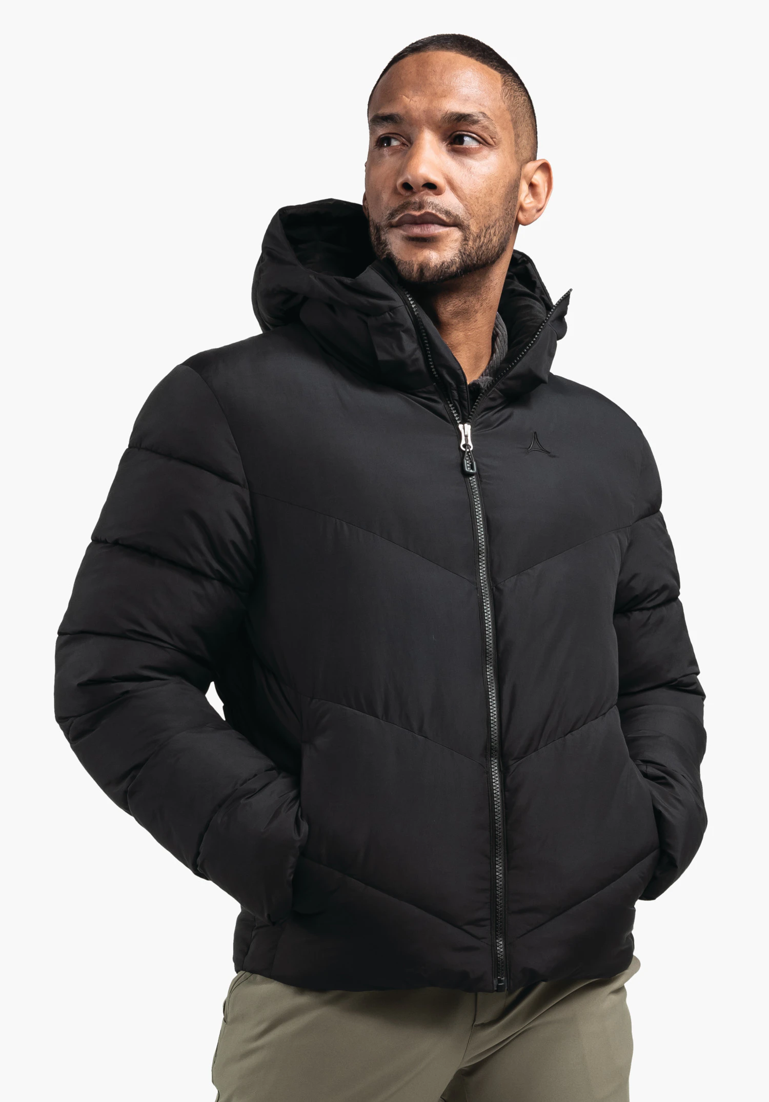 Ins Jacket Style Lodos MNS Schwarze Steppjacke mit Kapuze, von einem Mann getragen, Reißverschluss geschlossen, Hände in Taschen; Schöffel-Logo auf Brust; stehender Mann vor weißem Hintergrund.