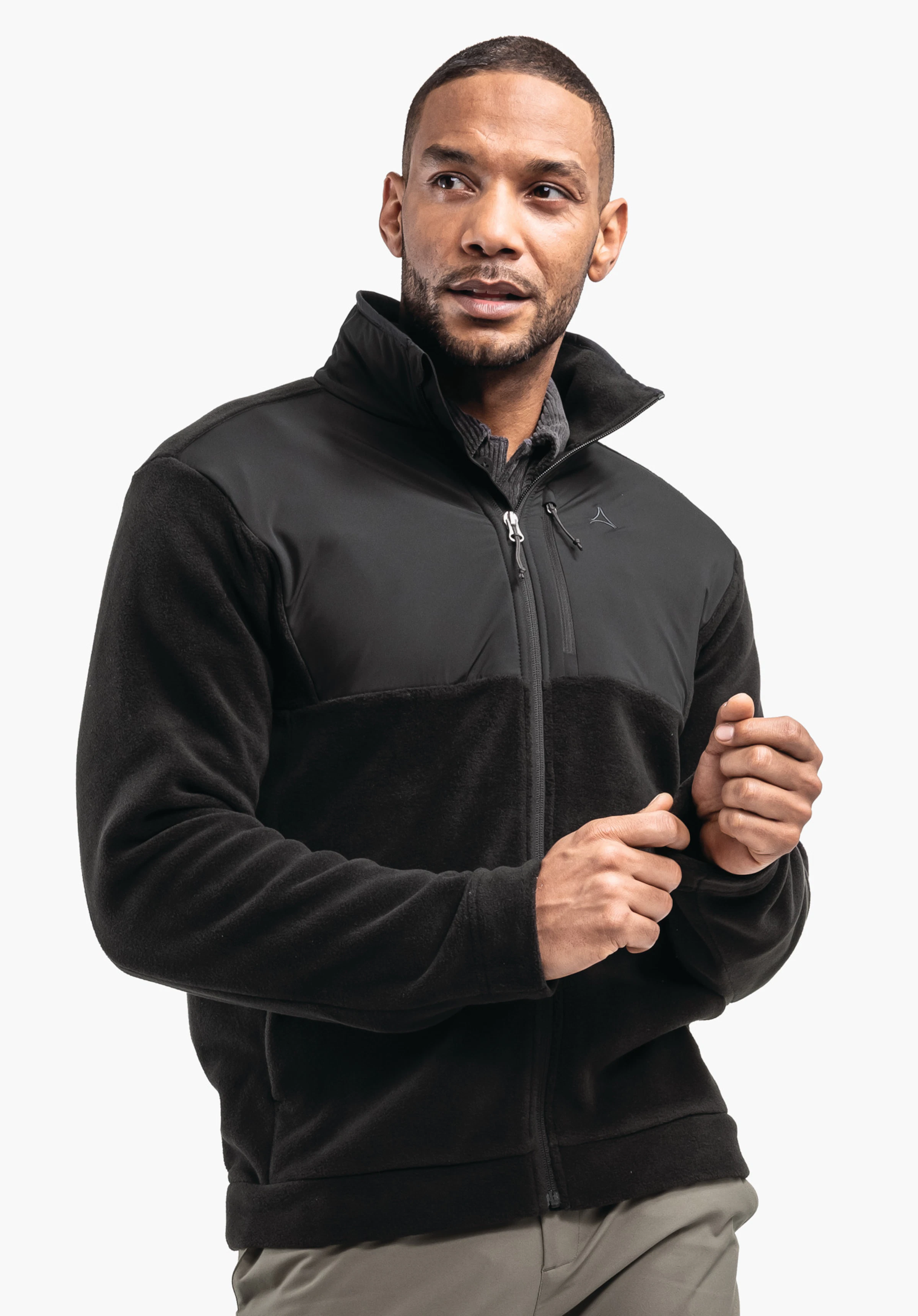 Fleece Jk Style Gregale MNS Homme portant une veste polaire noire avec logo Schöffel, tirant le zip; debout devant un fond blanc uni, pantalon gris clair.
