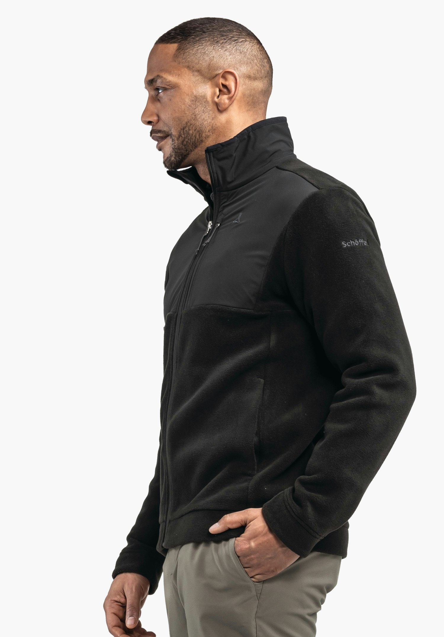 Fleece Jk Style Gregale MNS Schwarze Fleecejacke, Mann trägt sie seitlich stehend mit rechter Hand in Hosentasche vor hellem Hintergrund; Ärmelbeschriftung: "Schöffel"; kleines Schöffel-Logo auf Brust.