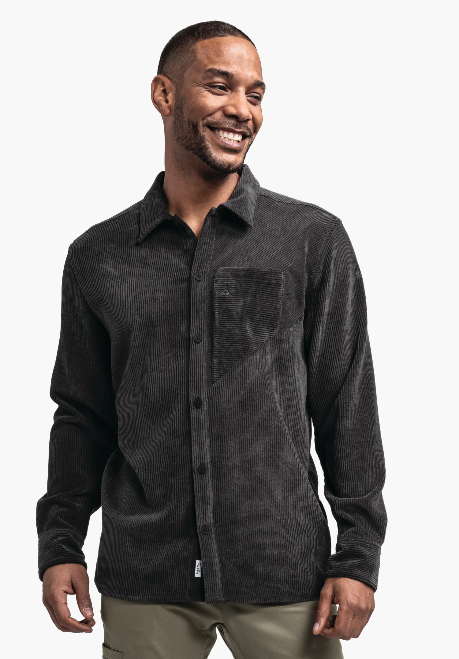 Shirt Style Lodos MNS Dunkel schwarzes Cordhemd mit Schöffel-Logo auf Brusttasche, getragen von lächelndem Mann, der nach rechts blickt; Knopfleiste sichtbar, hellgrüne Hose, weißer Hintergrund.