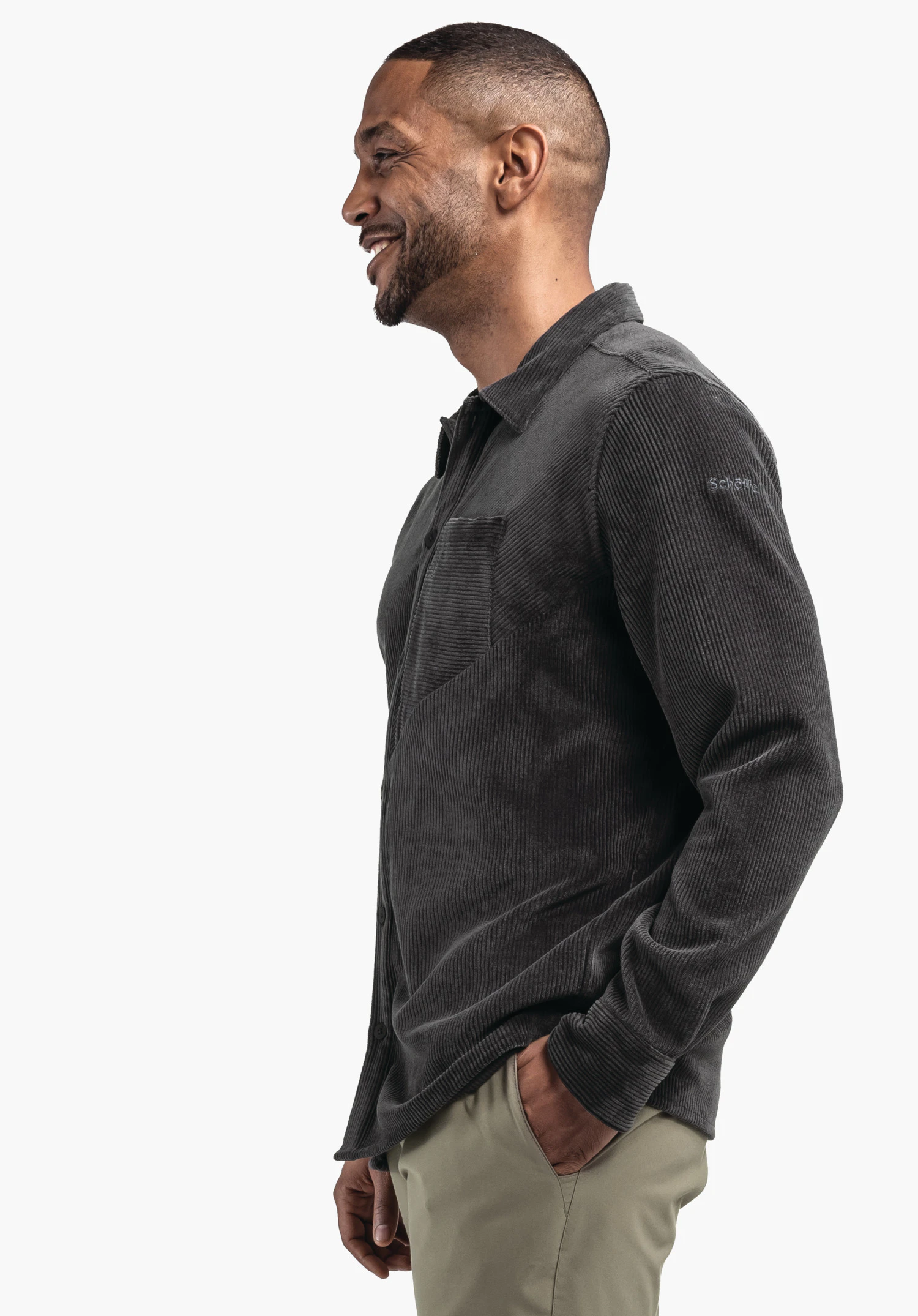 Shirt Style Lodos MNS Mann trägt dunkelgraues Cordhemd, lächelt und steht seitlich; Hand in Hosentasche, vor neutralem weißem Hintergrund. Text auf Ärmel: "Schöffel".