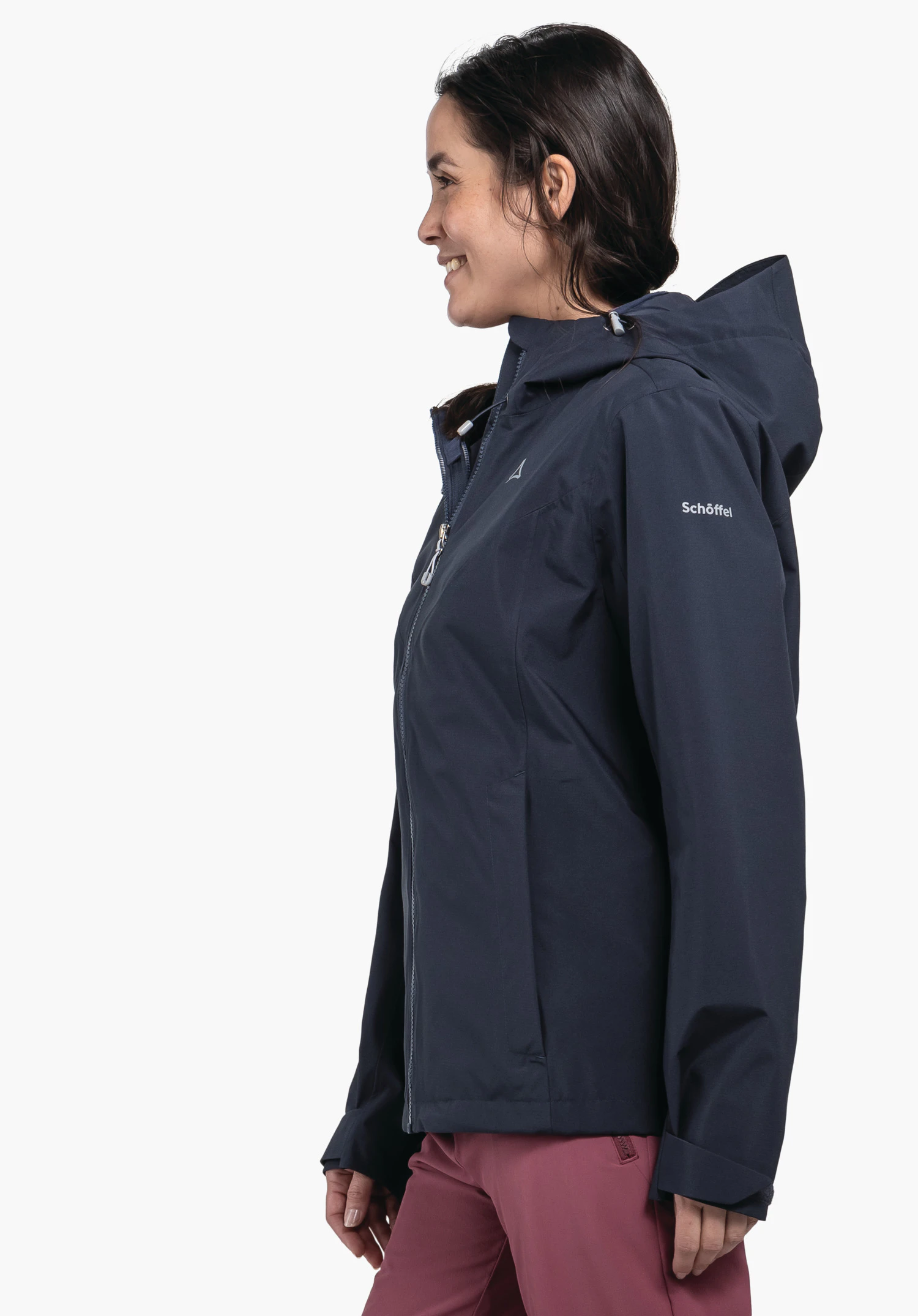 Jacket Style Yew WMS Frau trägt eine dunkelblaue Regenjacke mit Kapuze und dunkelroten Hosen, Schöffel-Logo auf der Brust, steht seitlich lächelnd vor neutralem weißem Hintergrund; Text: "Schöffel".