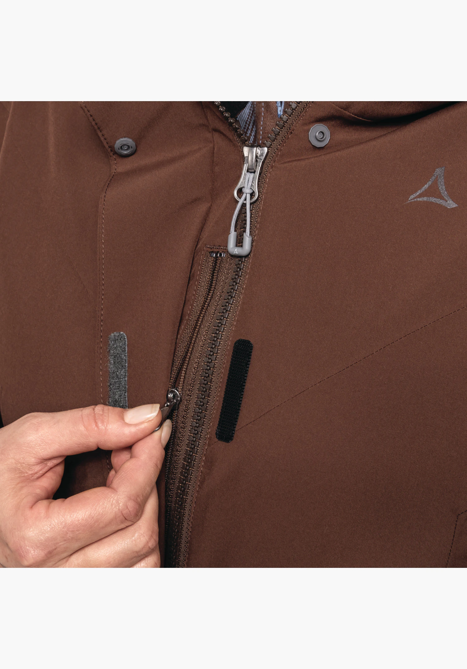 3in1 Jacket Style Tamina WMS zeigt den Frontreißverschluss mit Klettverschluss-Abdeckung, die von einer Hand gehalten wird, sowie das Schöffel-Logo auf dem braunen Material, Nahaufnahme vor neutralem Hintergrund.