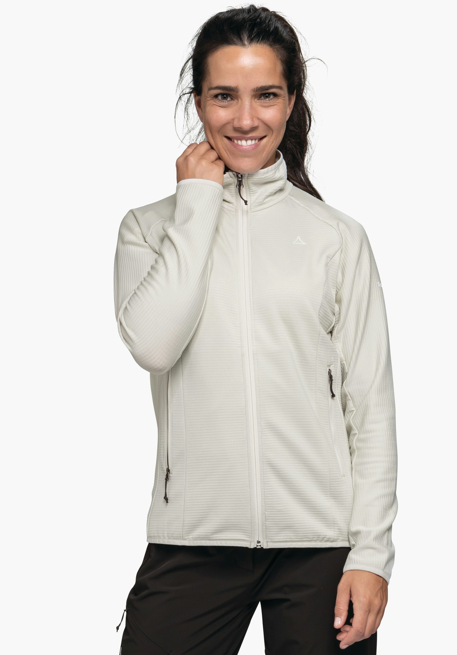 Fleece Jk Style Cascata WMS Veste portée par une femme en blanc cassé; elle sourit et tient le col avec la main droite; logo Schöffel sur la poitrine; pantalon noir, fond blanc.