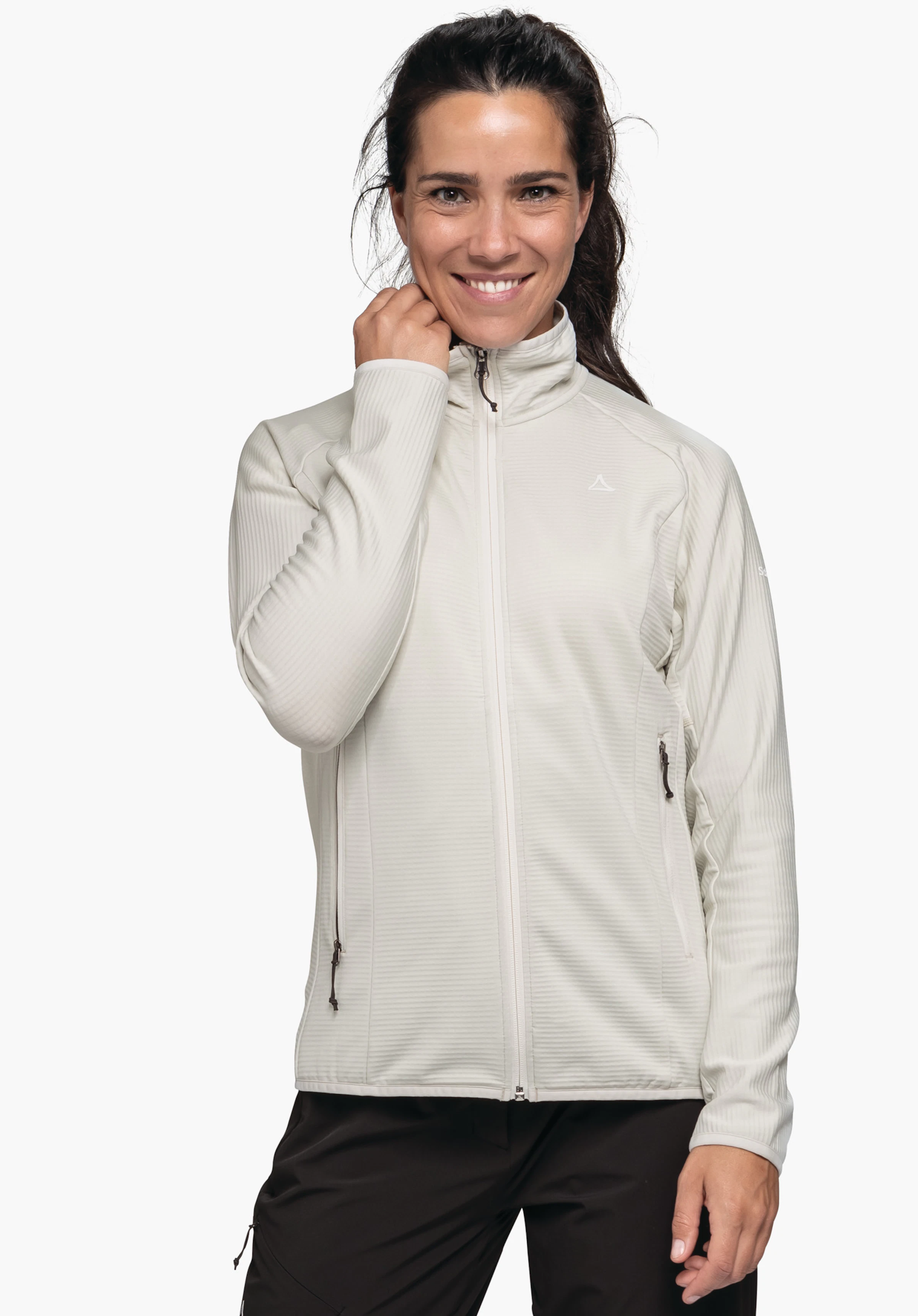 Fleece Jk Style Cascata WMS Veste portée par une femme en blanc cassé; elle sourit et tient le col avec la main droite; logo Schöffel sur la poitrine; pantalon noir, fond blanc.