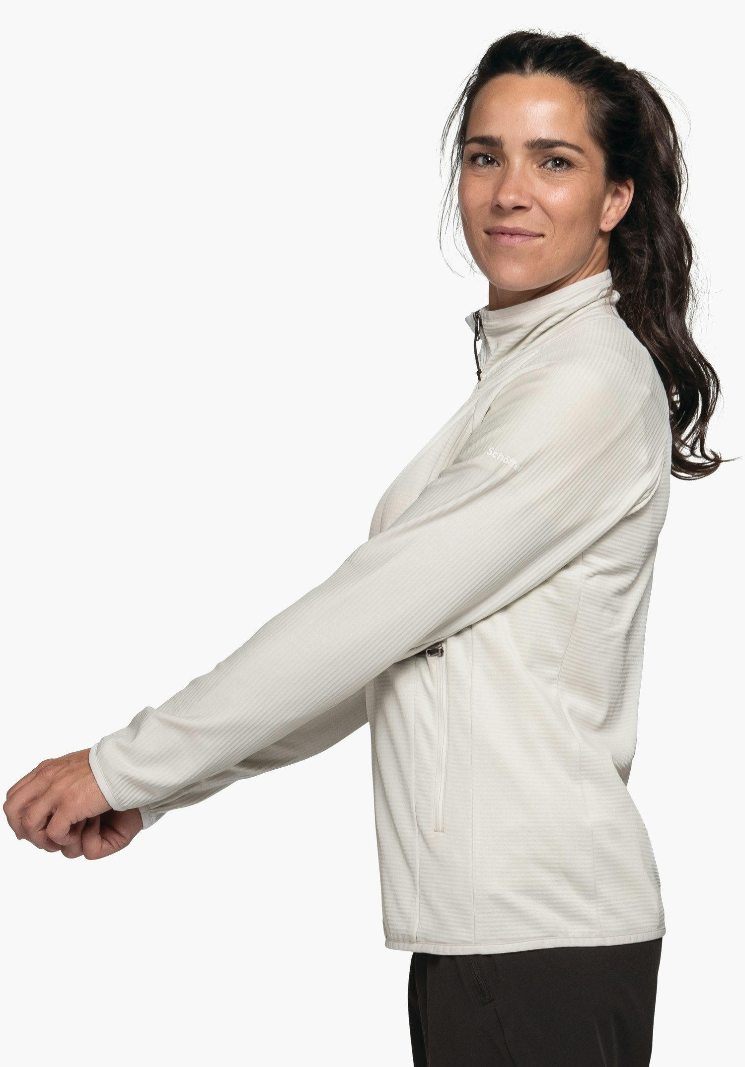 Fleece Jk Style Cascata WMS veste: femme porte une veste blanc clair à col montant, tend les bras vers l’avant; elle se tient de profil devant fond clair neutre, avec pantalon noir. Texte sur la manche: "Schöffel".