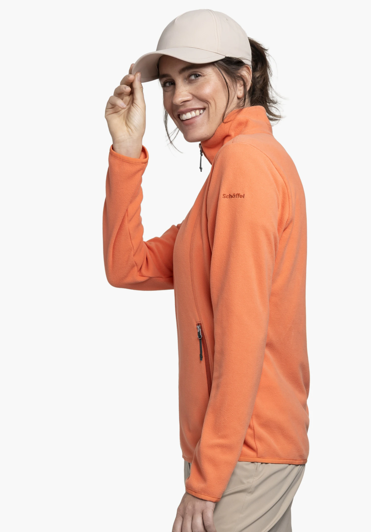 Fleece Jk Style Ash WMS in de kleur oranje. Het model staat zijwaarts voor een neutrale achtergrond en glimlacht terwijl ze haar pet bijstelt. Het subtiel geborduurde Schöffel-logo op de bovenarm benadrukt de hoogwaardige uitstraling van het fleecejack.