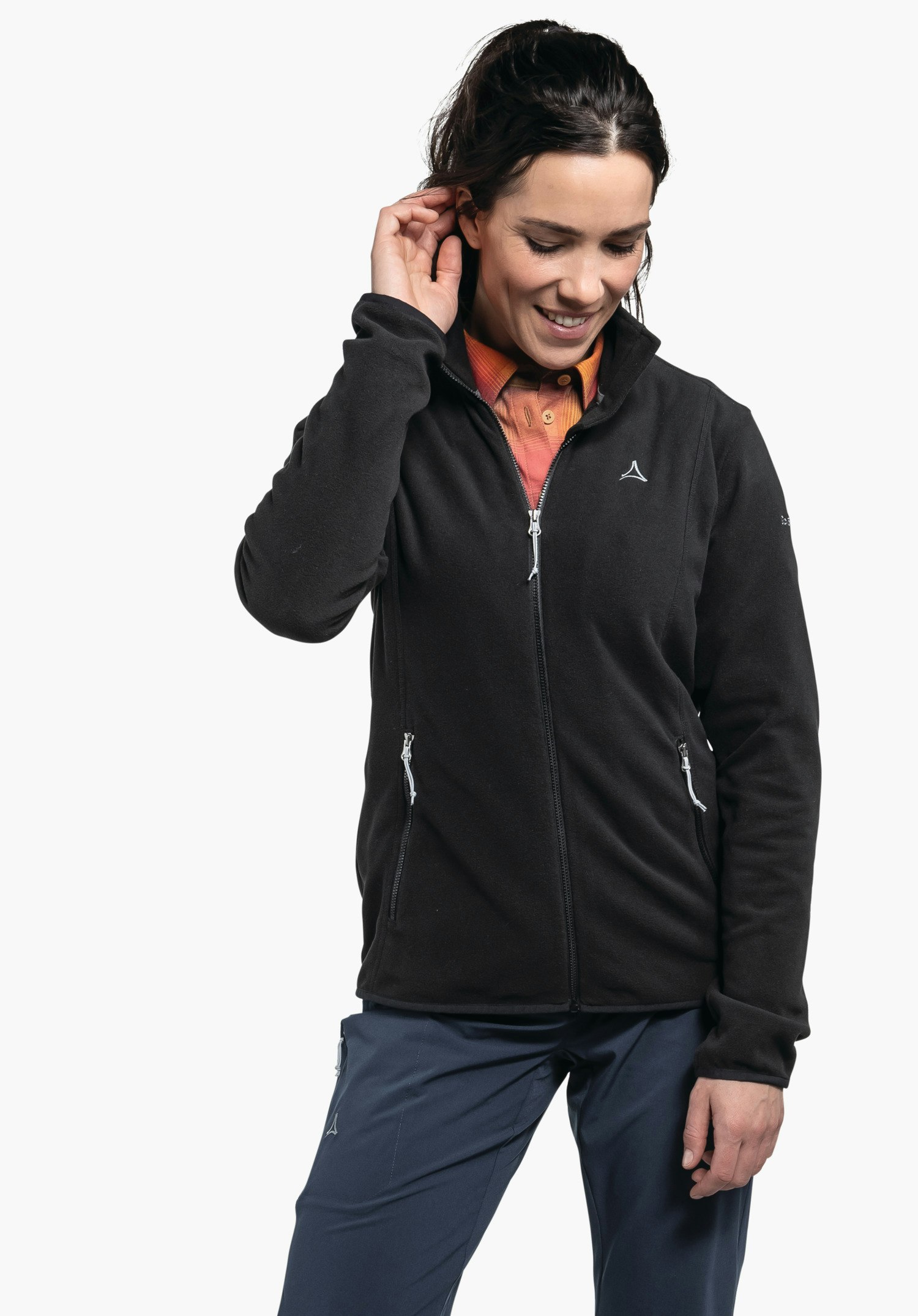 Fleece Jk Style Ash WMS Veste polaire noire avec zip et logo Schöffel, portée par une femme souriante touchant son oreille; chemise orange clair en dessous; pantalon bleu foncé; fond blanc.