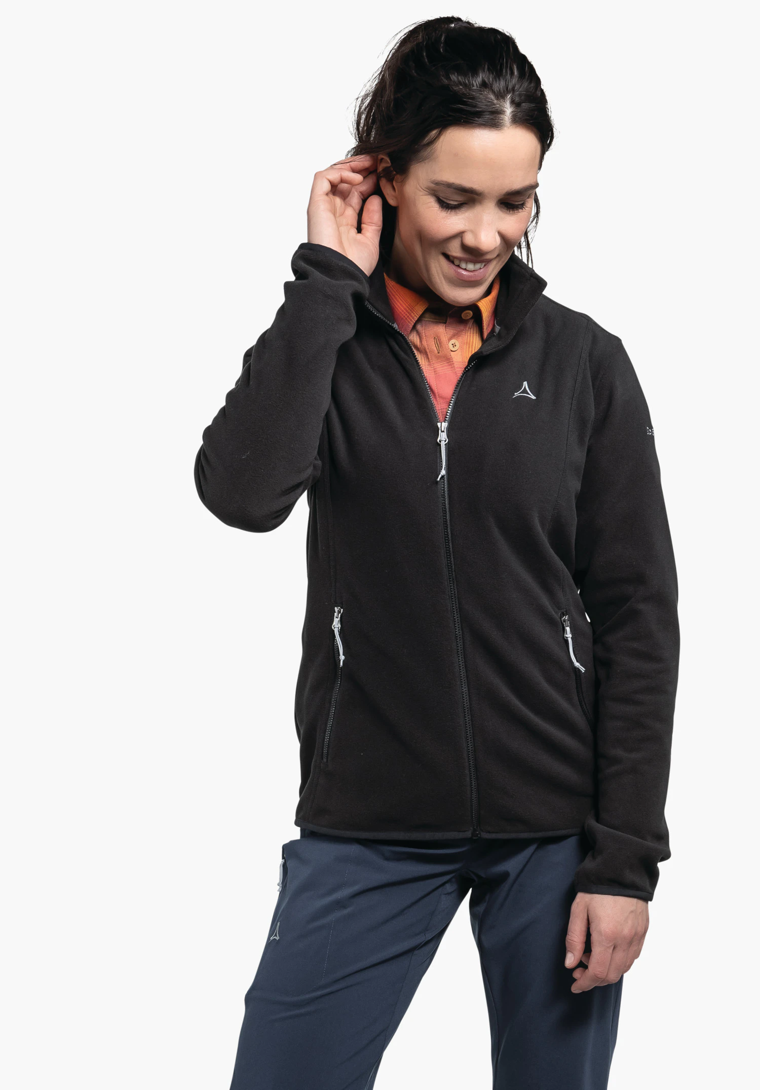 Fleece Jk Style Ash WMS zwarte fleecejas met ritssluiting en Schöffel-logo; gedragen door een lachende vrouw die haar oor aanraakt; lichtoranje overhemd eronder; donkerblauwe broek; witte achtergrond.