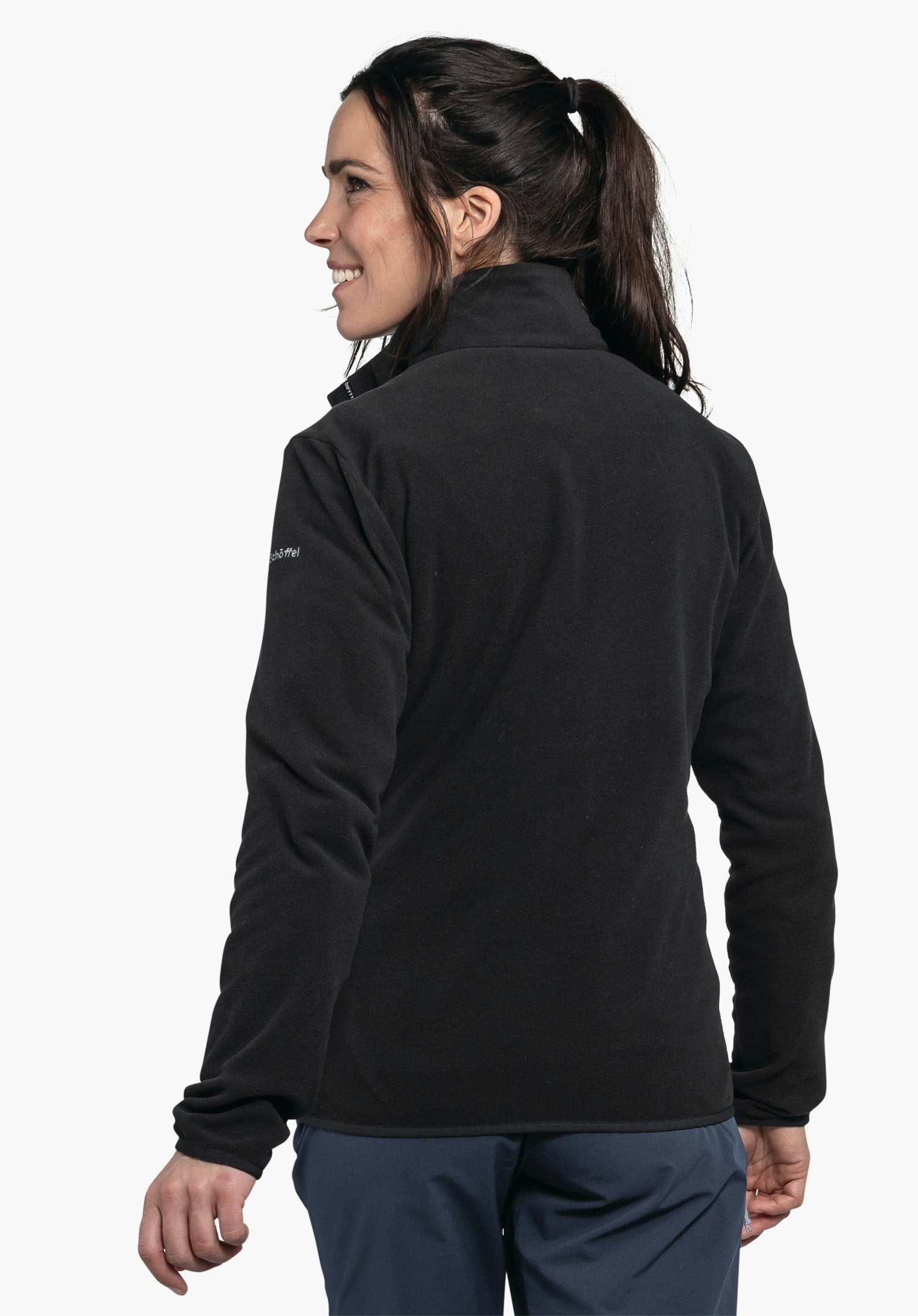Fleece Jk Style Ash WMS black fleece jacket worn by a woman in half profile, looking left; dark hair in a ponytail, dark blue trousers visible; neutral light background. Sleeve text: “Schöffel”.